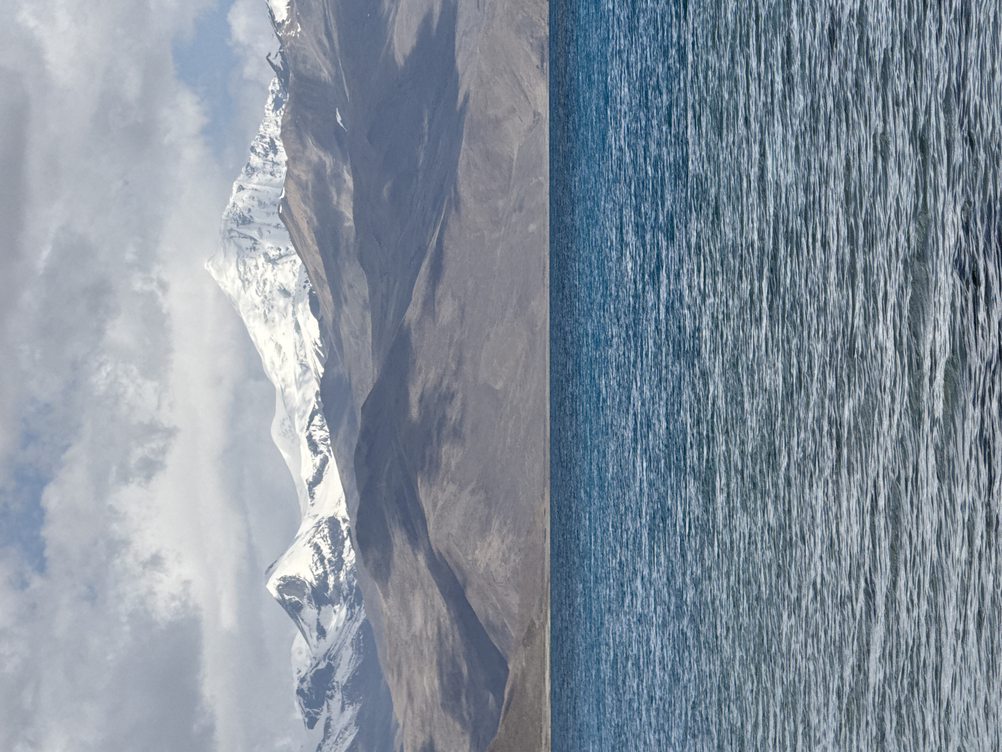 The image shows a landscape in Baidê, China. The left side features a mountain range with snow-capped peaks and rocky slopes under a cloudy sky. The right side is a body of water, appearing slightly rippled, and exhibiting a muted blue color. The image is bisected vertically.