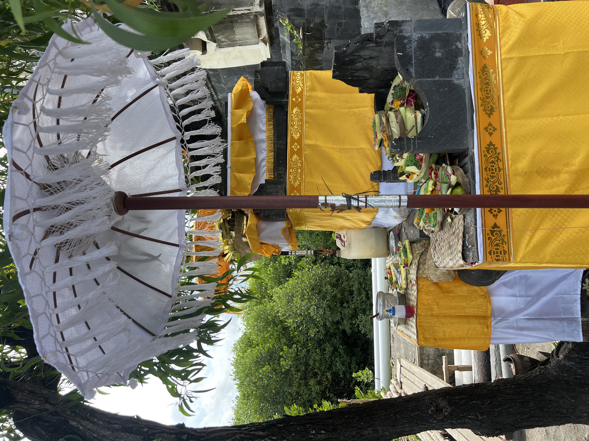 The image depicts a traditional Balinese shrine, possibly located in Denpasar, Indonesia, given the cultural artifacts and architectural style. The shrine features black tiered stonework partially draped in yellow fabric with gold trim and white cloth. Offerings, including woven baskets filled with greenery and flowers, are placed on the structure. A white umbrella with fringe stands beside the shrine. The setting includes a tree and foliage, suggesting an outdoor location. No people are visible.