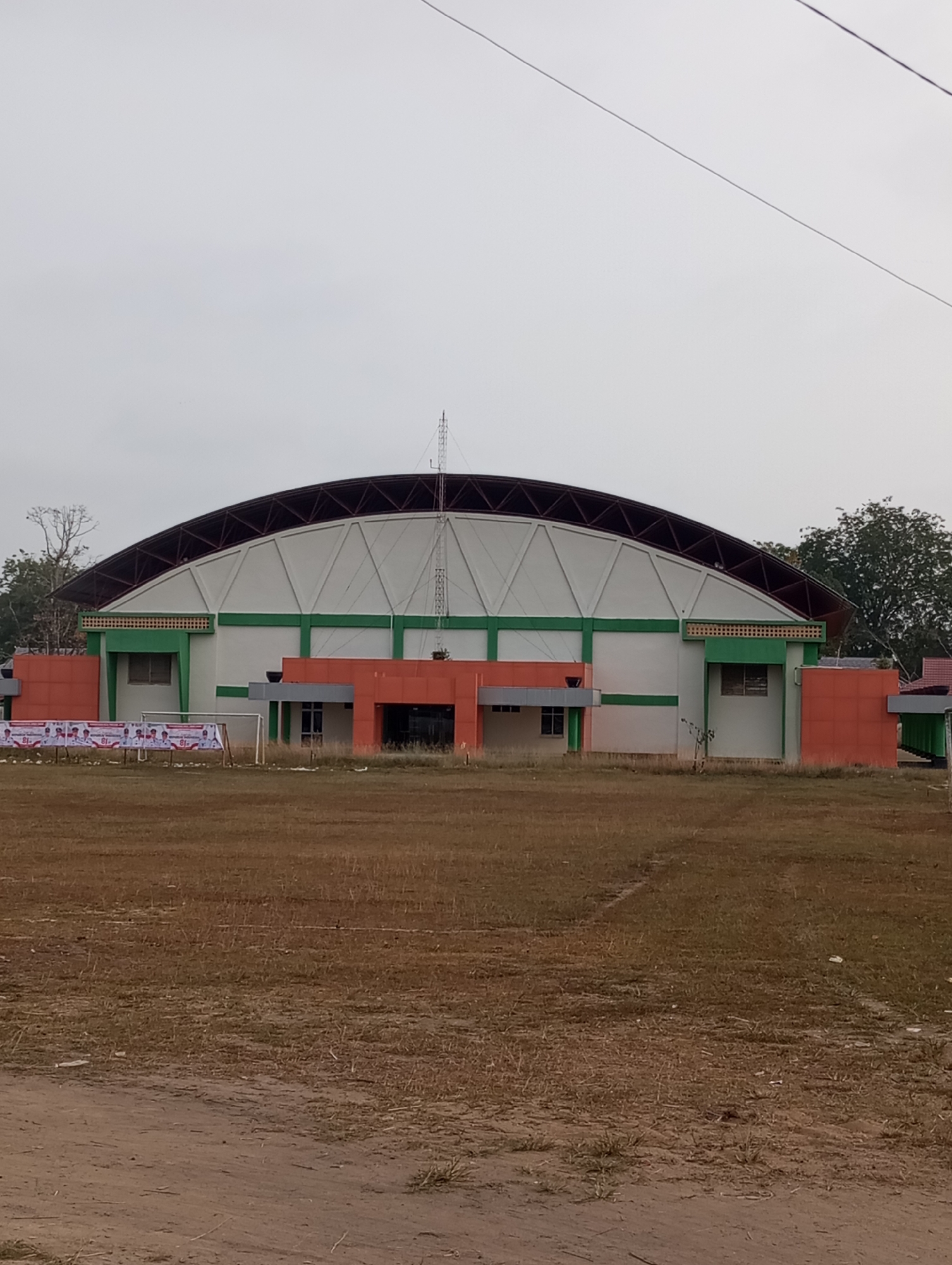 The image depicts an outdoor scene in Pematang Reba, Indonesia, featuring a large, modern sports arena with a distinctive arched roof. The arena's exterior is a combination of white and green sections, accented by orange paneling on the lower portions. Banners with Indonesian text and images are displayed in front of the building, suggesting an event or commemoration. A grass field, appearing somewhat dry and patchy, stretches out before the arena, with what looks like a dirt path in the foreground. Trees and foliage are visible in the background, with a clear, overcast sky overhead. There are no people or animals visible in the frame.