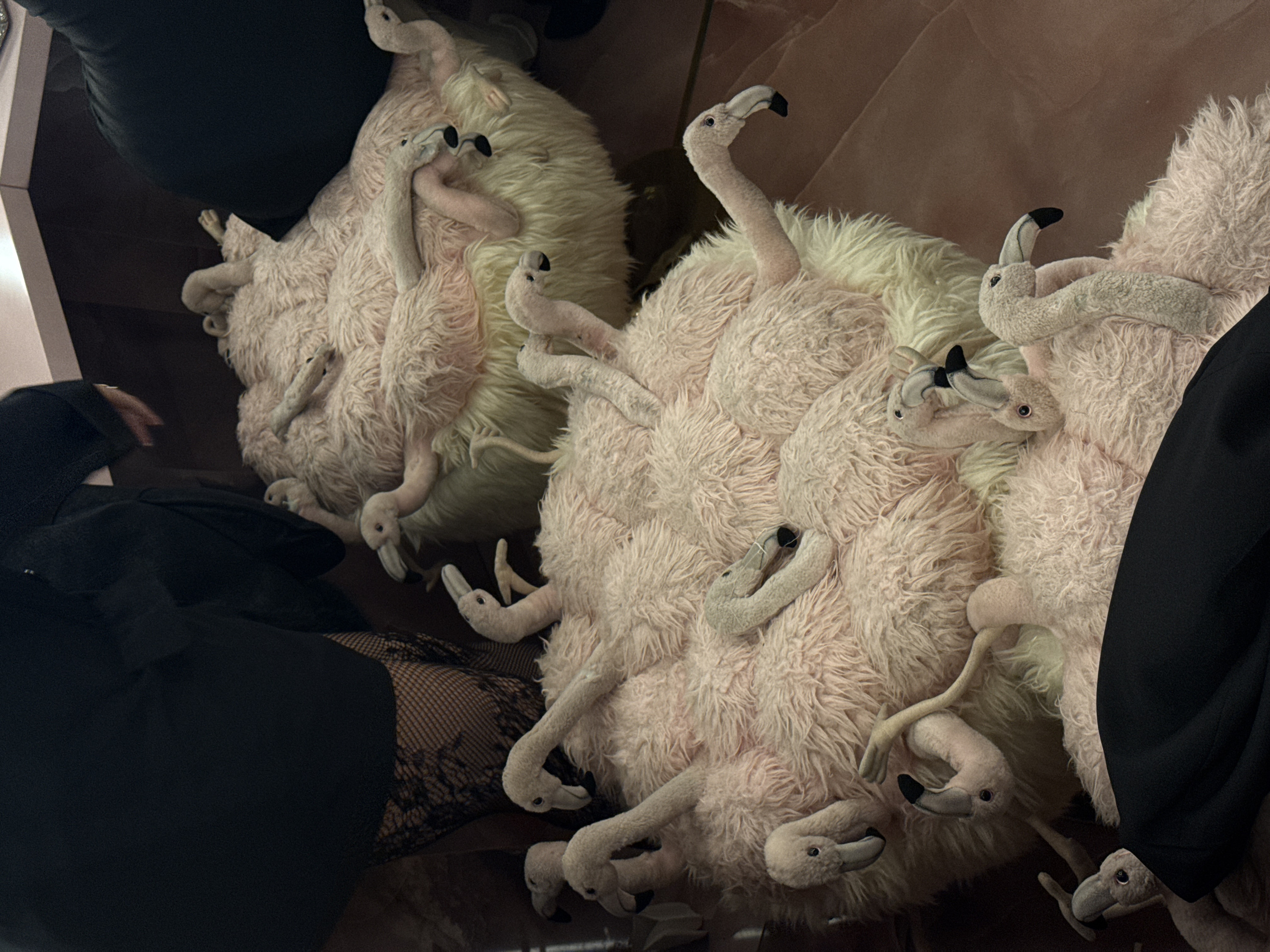 The scene is an indoor environment in Milan, Italy, characterized by dim, artificial lighting. The foreground is dominated by several plush, stylized flamingo objects. Two large, roughly spherical or oval forms serve as primary subjects, upholstered in shaggy light pink and cream-colored faux fur. Embedded within these forms are numerous smaller, plush flamingo heads and necks, rendered in light pink fabric with black-tipped beaks and small dark eyes. Additional individual plush flamingo heads, necks, and some leg elements are scattered among and around these larger structures.
To the upper left, the lower torso and legs of an individual are partially visible, clad in dark, pleated fabric over black patterned hosiery. A light-skinned hand rests upon a light-colored, flat horizontal surface, likely a countertop. A segment of dark, plain fabric, indicative of clothing, is visible at the bottom right. The background features a smooth, light-colored horizontal counter or shelf situated above a smooth, dark reddish-brown vertical wall or panel. The floor is a dark, indistinct surface.