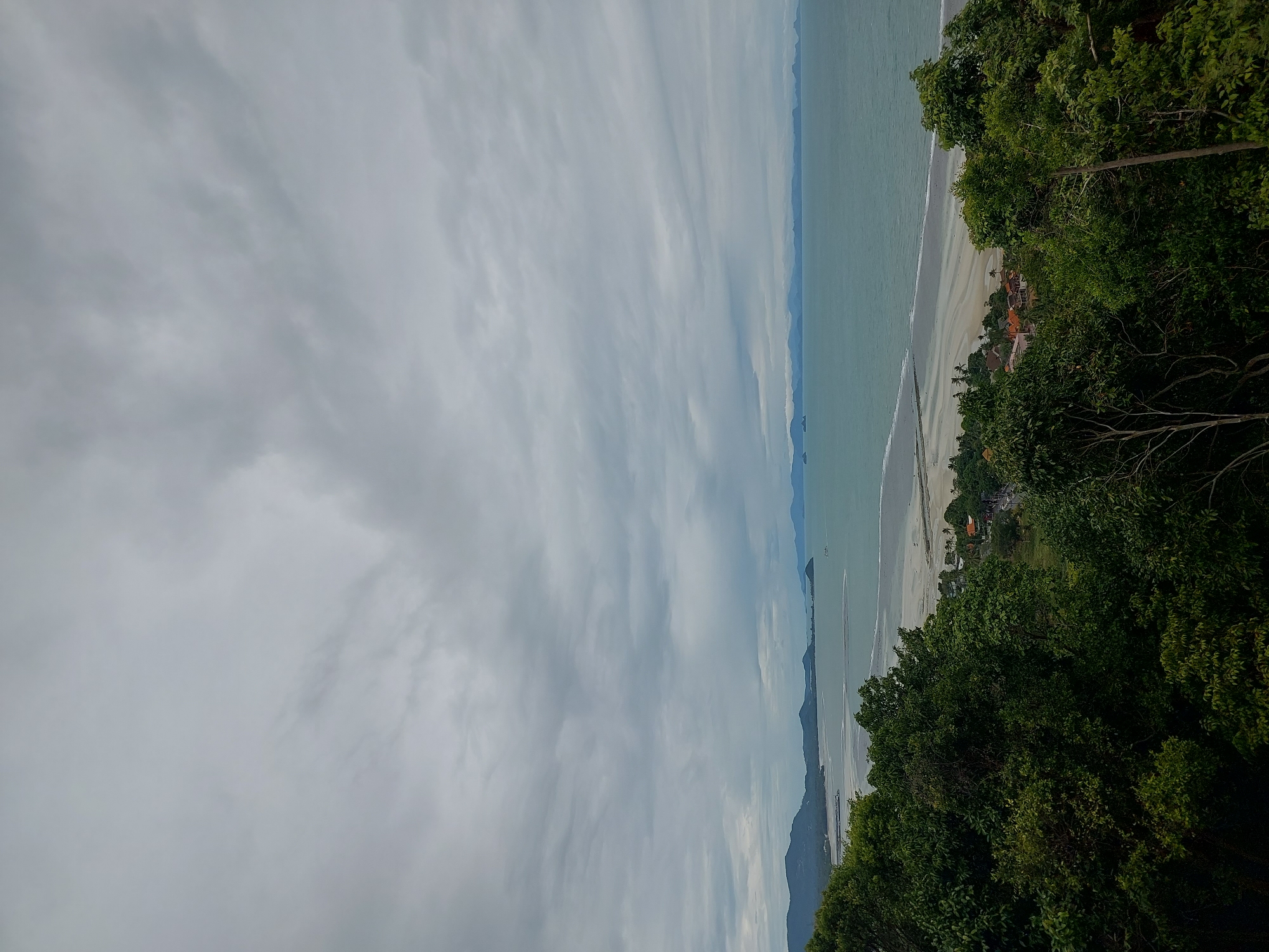 An elevated, wide-angle, full shot captures a panoramic view of the coast of Ko Samui District, Thailand, under an overcast sky. In the foreground, lush green trees fill the bottom half of the frame, obscuring a view of the terrain below. The middle ground reveals a sandy beach bordering a calm, turquoise ocean. A small cluster of buildings with orange roofs is visible near the shore, with a road winding through the area. In the distance, a chain of mountainous islands stretches across the horizon, fading into the misty sky. The sea is mostly calm, with a faint trail of white from a distant boat. The overall mood is serene, with the overcast sky contributing to a soft, diffused light.