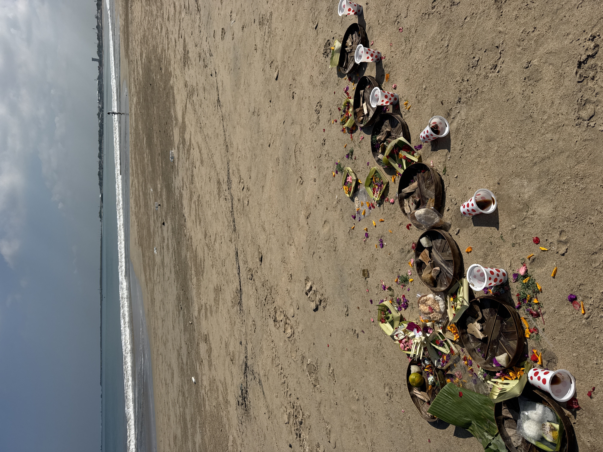 This image captures a serene scene on a sandy beach in Kuta Selatan, Indonesia, depicting a traditional Balinese Hindu offering ceremony.
The foreground and midground feature a line of offerings arranged on the light brown sand, stretching from the lower right towards the upper right of the frame. These offerings consist primarily of numerous small, circular woven bamboo or straw containers, reminiscent of *canang sari* trays. Many of these contain vibrant offerings of colorful flowers (red, yellow, and purple petals), green leaves (including banana leaf segments), and small food items like fruit or what appears to be puffed rice. Scattered loose flower petals are also visible on the sand around the offerings. Interspersed among these are several white paper cups with red polka dots, some of which contain a dark liquid, likely an offering.
To the left, the calm, greyish-blue ocean stretches to the horizon, with gentle white waves breaking on the shore. A distant coastline is visible, showing faint silhouettes of buildings or structures under a partly cloudy sky. A solitary, small figure can be seen standing near the water's edge in the far distance.
The scene suggests a daytime setting, with soft, ambient lighting. There is no visible text in the image. The overall impression is one of spiritual devotion and cultural tradition taking place in a natural coastal environment.