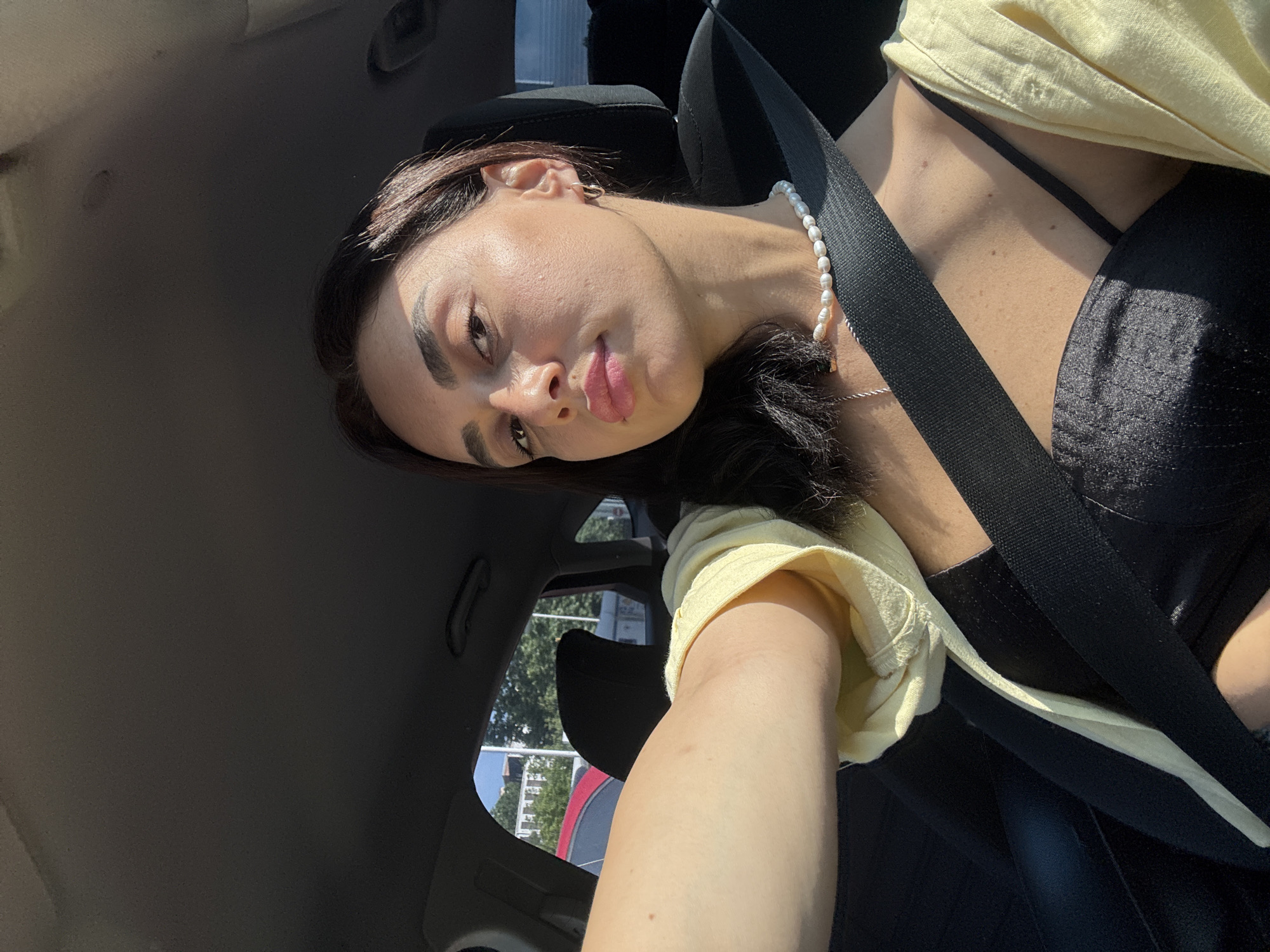 In a sunlit selfie taken from inside a car, a young woman with dark hair and prominent cheekbones is captured, wearing a black top and a light yellow open shirt. She has a pearl necklace adorning her neck and small hoop earrings. The woman is looking directly at the camera with a neutral expression, her lips slightly parted. Sunlight casts strong shadows across her face, highlighting her features. The background shows the interior of a car, including a seatbelt across her chest and a glimpse of trees and buildings through the car window, suggesting an urban environment. The scene appears to be in Tbilisi, Georgia, given the architectural style visible outside the car.