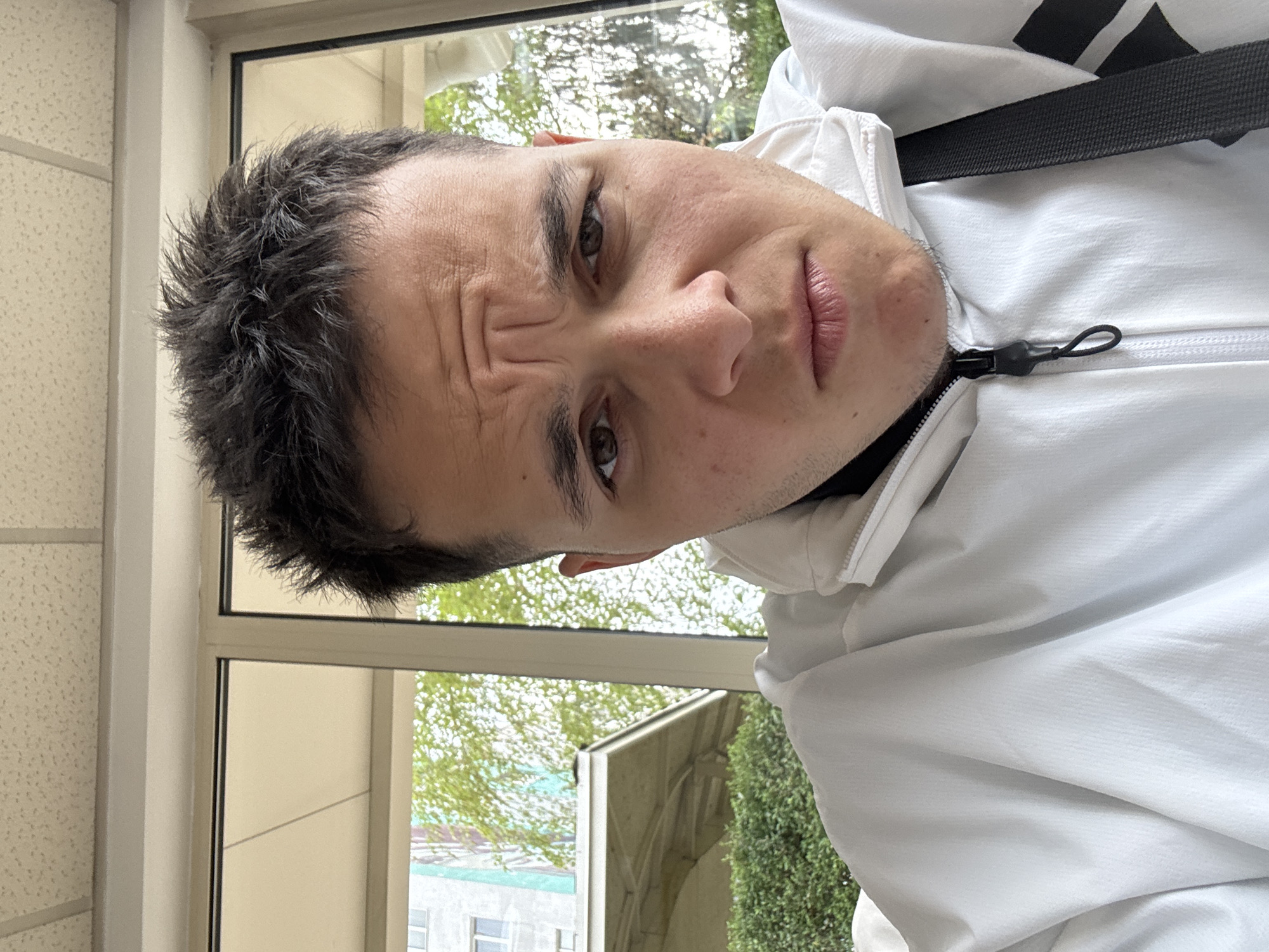 A medium close-up, tilted portrait features a young man with dark, spiky hair and expressive dark eyes, looking directly at the camera. He wears a white hooded jacket with a black zipper and a black strap, possibly from a bag, visible across his chest. His eyebrows are furrowed, creating prominent horizontal wrinkles on his forehead, and his lips are slightly pursed, conveying a skeptical, questioning, or mildly contemplative expression. A faint stubble is visible on his chin and jawline.
The man is positioned indoors, adjacent to a large window that reveals a bright, daytime view. Outside, lush green trees dominate the mid-ground, suggesting an outdoor garden or park area, with parts of light-colored buildings visible further in the distance. The interior setting includes a light-colored wall or ceiling with a textured, possibly acoustic, surface on the left side of the frame, and a clear window frame. The natural light filtering through the window illuminates the scene. The image itself is taken at a significant diagonal tilt, orienting the man's head towards the upper right corner of the frame. There is no visible text in the image. The location is Sofia, Bulgaria.