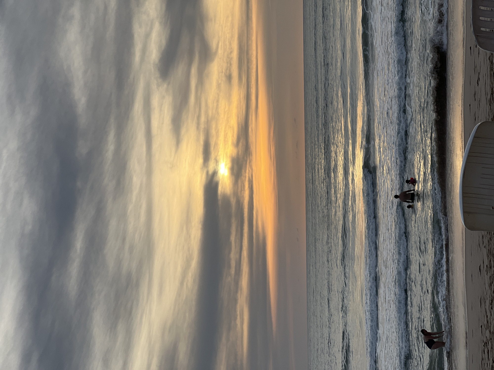 The image depicts a beach scene at dusk in Choeng Thale, Thailand. The sky is filled with dramatic, striated clouds in shades of gray and pale yellow, with the sun partially obscured by clouds, casting a warm, golden light on the horizon. The ocean is calm with gentle waves breaking on the shore. In the foreground, a sandy beach is visible, with two white chairs facing away from the camera. In the water, several people are present: a person in a black swimsuit is bending down near the shore, another person is standing in the water with two smaller figures, and a child in a red top is further out in the water. The overall atmosphere is tranquil and serene, indicative of a peaceful evening by the sea.