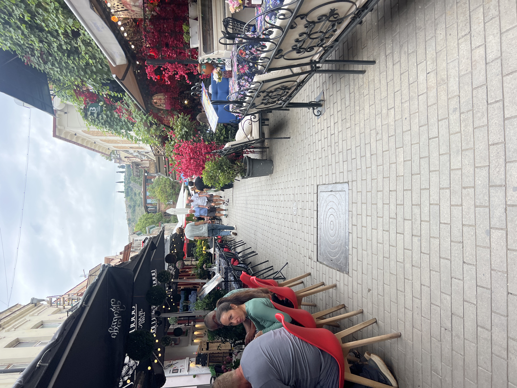 The image depicts a view of a pedestrian street in Tbilisi, Georgia, likely in the Old Town or a similar historic district, characterized by buildings with balconies and lush greenery. The street is paved with light-colored rectangular bricks. On the left, several outdoor dining areas are set up with black umbrellas bearing Georgian script and white lettering, some decorated with hanging spherical topiaries and string lights. Red chairs are visible at some tables. On the right, a restaurant or cafe features abundant red bougainvillea flowers cascading down the building's facade, along with outdoor seating. Diners are seated at tables on both sides of the street, and several people are walking along the pavement. The overall atmosphere suggests a vibrant, popular dining and strolling area.
