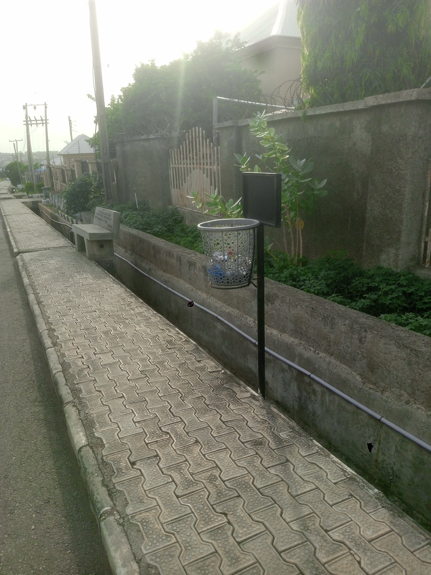 This outdoor image from Kubwa, Nigeria, depicts a paved pedestrian walkway alongside a concrete drainage ditch, leading into a residential area under bright, hazy sunlight. The time of day appears to be either morning or late afternoon due to the strong, low-angle light causing lens flare at the top right.
**Subjects & Objects:**
In the foreground, a patterned grey concrete paver walkway extends into the distance, bordered on the left by an asphalt road. A concrete drainage ditch runs parallel to the walkway, containing a white PVC pipe along its inner wall. A silver, perforated metal trash can, mounted on a black pole, stands next to the ditch and contains some blue and white plastic refuse. Further along the walkway, a plain grey concrete bench with a backrest is visible. Beyond the ditch, a concrete boundary wall topped with barbed wire separates the path from green foliage and trees. Parts of residential buildings, including a house with a grey, sloped roof, are discernible through the trees. Utility poles and electrical wires line the street in the background.
**Activity or Event:**
No human activity or specific event is taking place; the scene is static.
**Notable Details:**
The concrete bench features inscribed text on its backrest, reading: "THE NEW KUBWA RESIDENTS", "DEVELOPMENT ASSOCIATION", "A NEW DAWN FOR KUBWA", "BEAUTIFICATION & CLEANLINESS", and "YOUR PARTNER IN PROGRESS". The overall impression is a calm, maintained suburban environment.