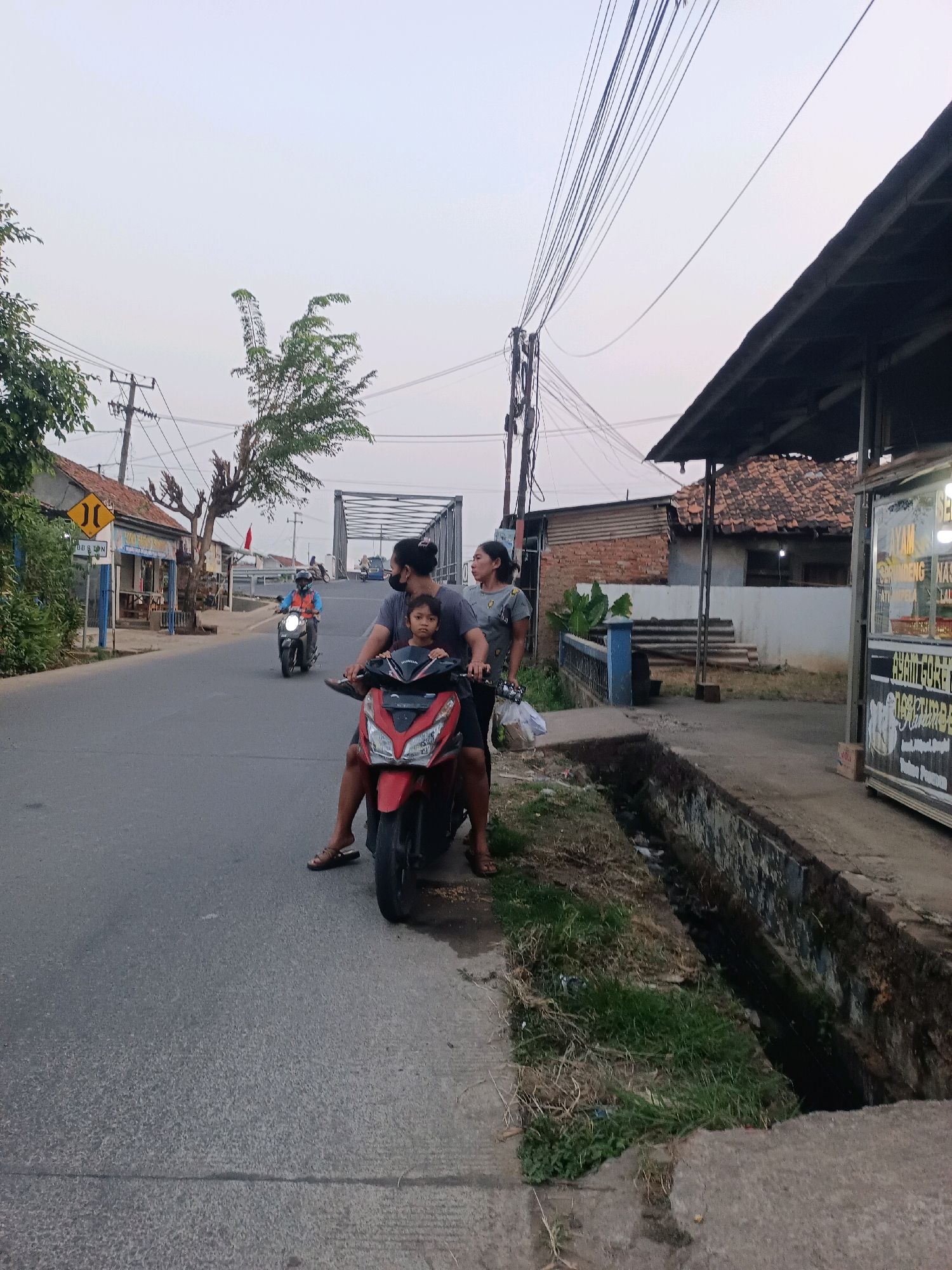 In Karawang, Indonesia, a family of three is riding a red motorcycle on a paved road. A man is driving, a woman is sitting behind him, and a young child is in front of the man. They are all looking in the same direction, presumably forward. The road is lined with a drainage ditch on the right and vegetation. In the background, there are buildings, utility poles with many wires, and a bridge. Another motorcycle with a rider wearing a blue vest is visible further down the road. The sky is overcast.