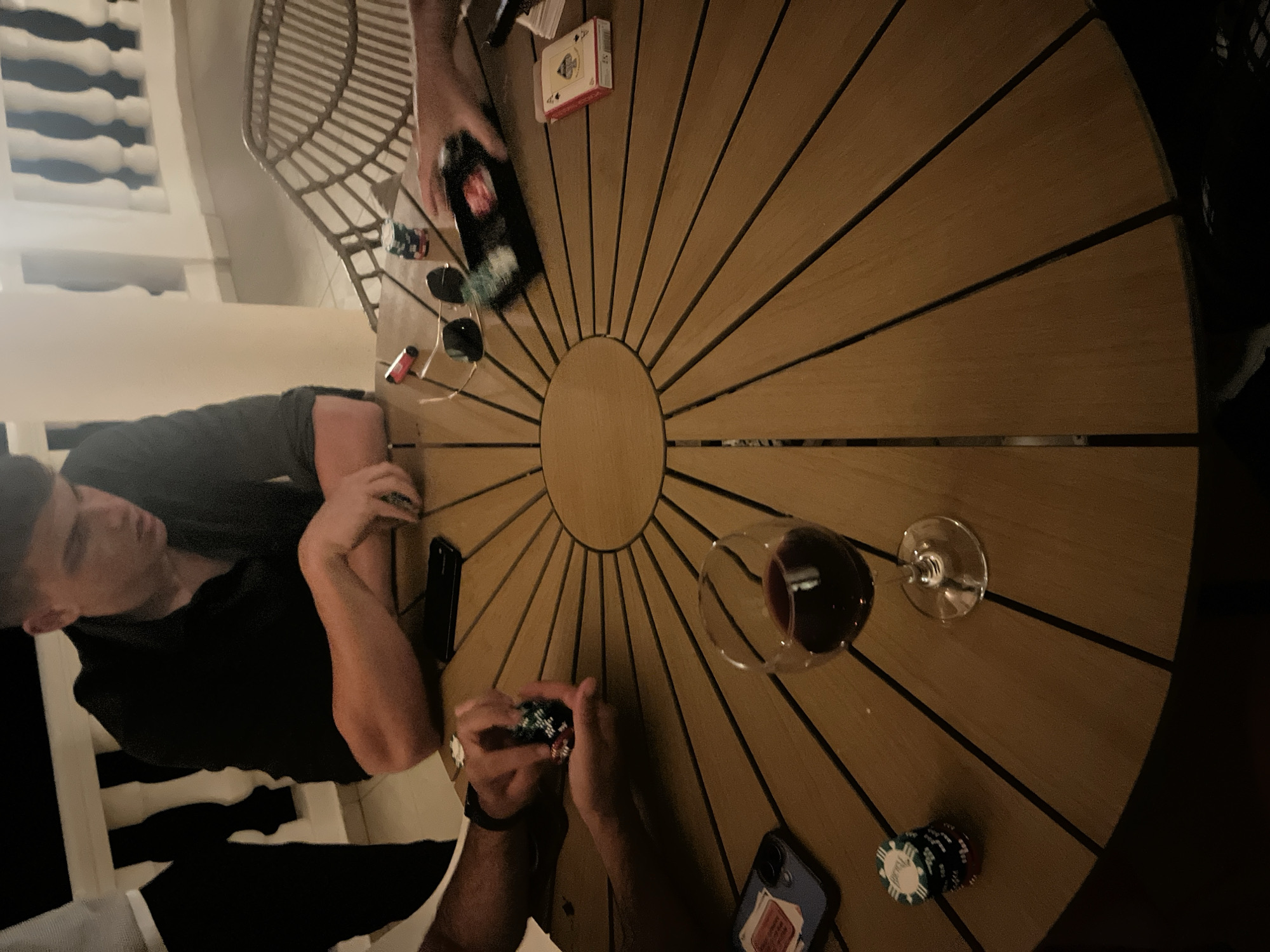 Captured from an elevated angle, the image portrays a social gathering in el Poble Nou de Benitatxell / Benitachell, Spain. A group of people are seated around a circular wooden table with a sunburst pattern, engaged in a poker game. Objects on the table include playing cards, poker chips, smartphones, a bottle of liquor, sunglasses, a lighter, and a glass of red wine. At least three individuals are partially visible: one with his arms resting on the table, and two others whose hands are visible, one of whom is holding poker chips. The surrounding environment includes a wicker chair and a white balustrade. The scene suggests an evening or nighttime setting, possibly on a patio or terrace.