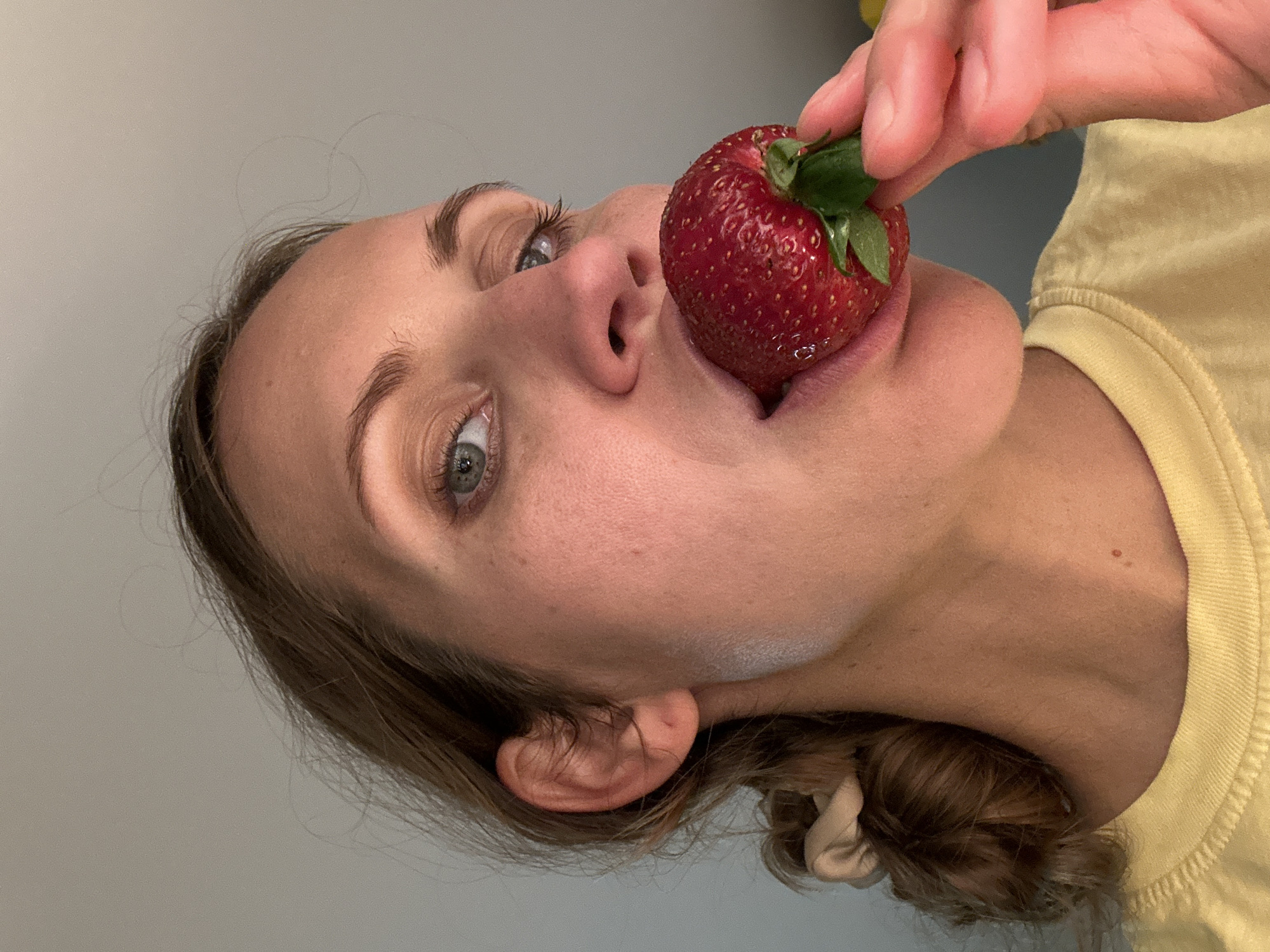 A light-skinned woman with brown hair pulled back in a ponytail, wearing a yellow shirt, is holding a large strawberry to her mouth. The strawberry is vibrant red with green leaves and appears to be the focal point. The background consists of a plain gray wall. The location cannot be determined from the image.