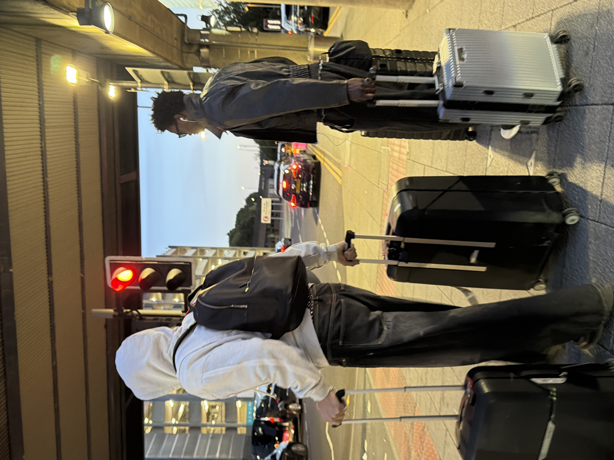 Two individuals are standing on a sidewalk next to luggage at a street intersection in Crawley, United Kingdom, with a traffic light showing red. The person on the left, facing away from the camera, wears a white hooded sweatshirt, a black backpack, and dark jeans. They are holding the handle of a black rolling suitcase. To their right, a second individual, also facing away from the camera, is wearing a dark jacket, dark pants, and white earbuds, and glasses. They are holding the handle of a silver rolling suitcase positioned behind a larger black rolling suitcase. In the background, cars are stopped at the intersection, and a multi-story car park structure is visible, illuminated by lights. The sky is a dusky blue, indicating evening or early morning.