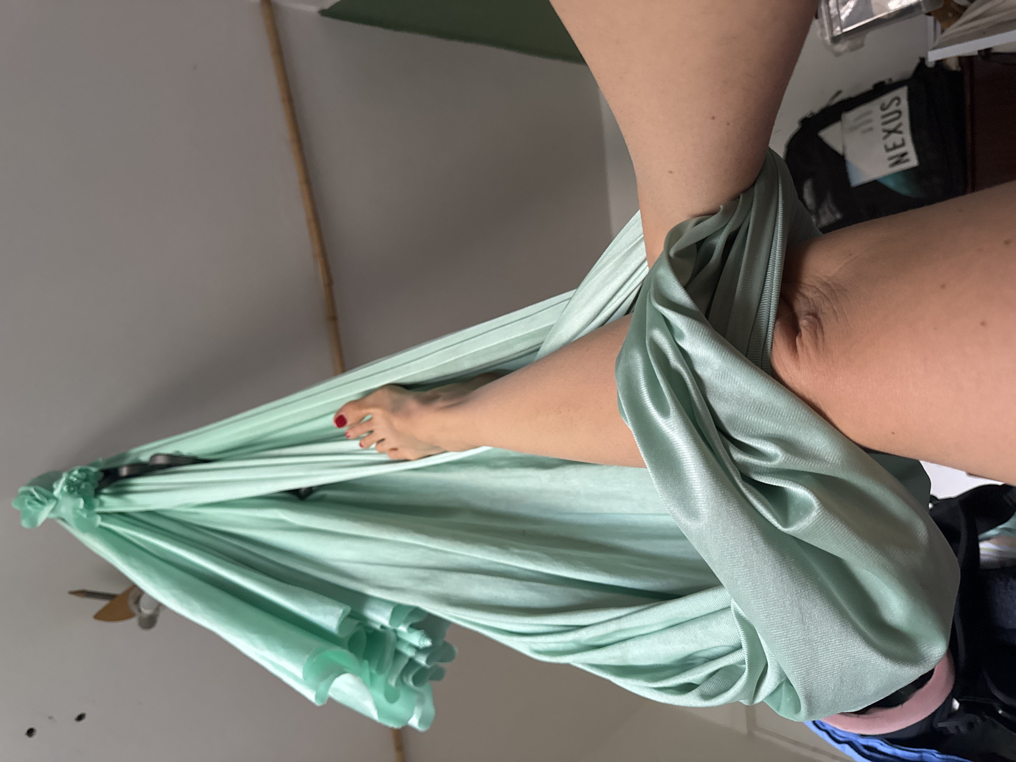 The image shows a person's legs suspended in a light green aerial hammock. One foot, with red-painted toenails, is visible. The hammock is made of a silky material that drapes and folds around the legs. The background is a plain wall, with a hint of a green ceiling and a wooden pole. To the right, a black bag with "NEXUS" written on it is partially visible, along with other objects suggesting an indoor environment. The lighting appears to be natural, coming from an unseen source. The scene is static and focuses on the interaction between the person and the aerial hammock.