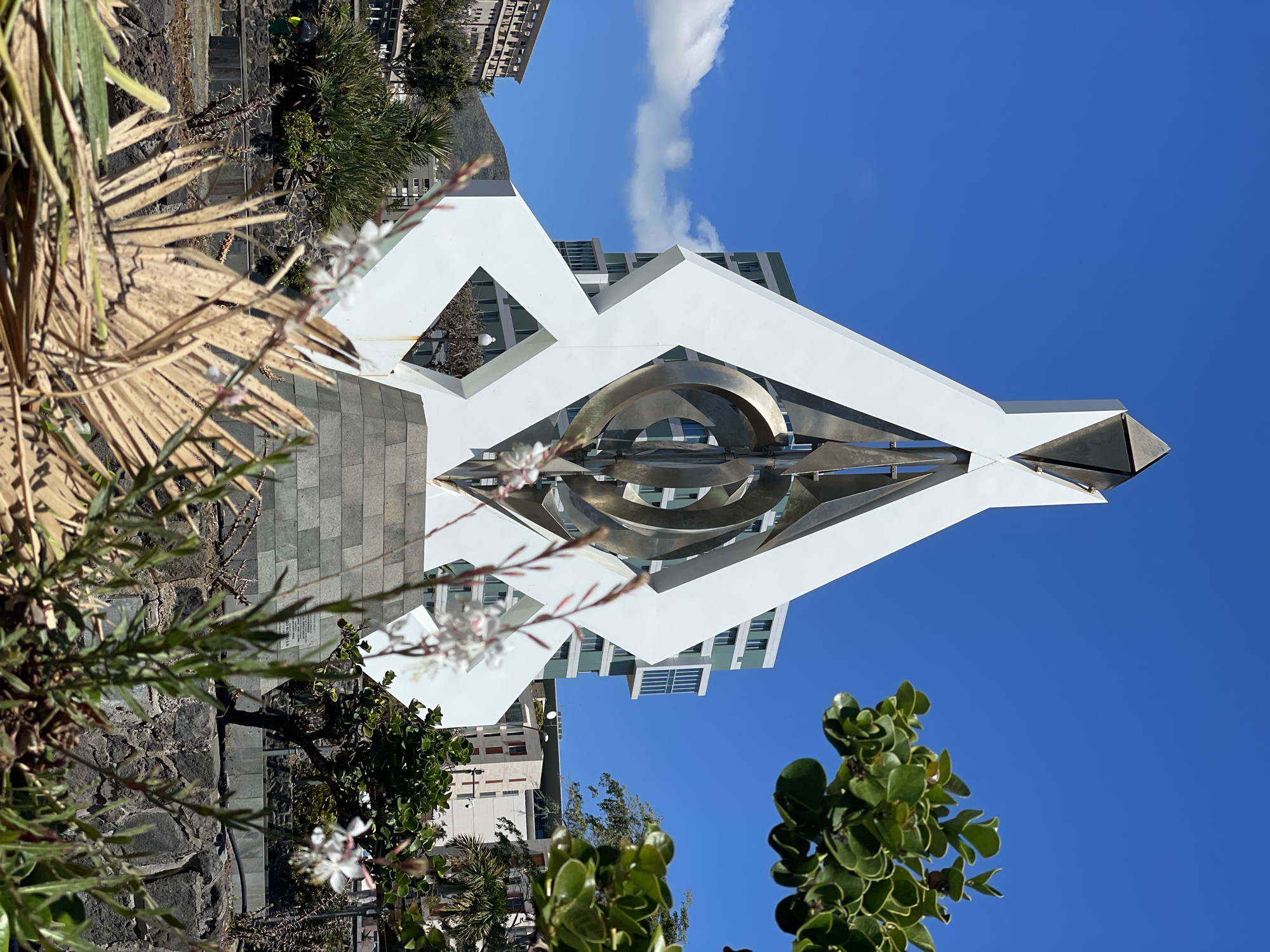 The image depicts the "Plaza de España" monument in Santa Cruz de Tenerife, Spain. The monument is a prominent white structure with geometric shapes and a central metal sculpture composed of concentric circles. In the background, buildings and the sky are visible, with a few scattered clouds. In the foreground, there is vegetation, including trees, bushes, and flowers. The image appears to be taken from a slightly low angle, looking upwards towards the monument. The lighting suggests a sunny day, with clear blue skies.