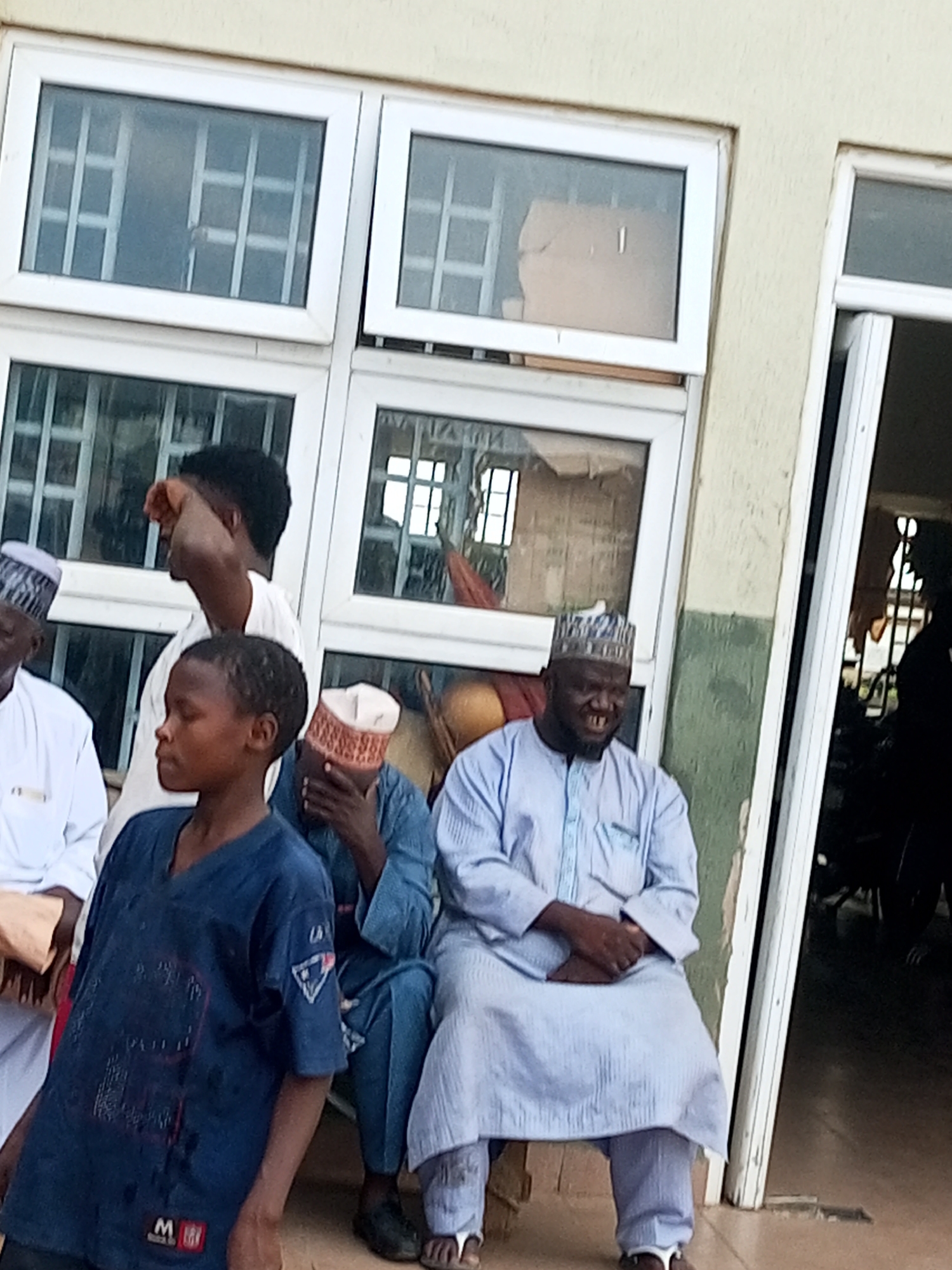The image shows a group of people outside a building with large white-framed windows. The building appears to be an institutional structure, possibly a school or administrative office, in Gombe, Nigeria. In the foreground, a young boy in a dark blue t-shirt stands looking to his left. Behind him, several men are seated and standing. One man, wearing a light blue traditional outfit and a patterned cap, is seated and smiling, showing his teeth. Another man in a blue garment and a red and white patterned cap is partially visible behind the boy. To the left, a man in a white garment and a patterned cap is standing, looking away from the camera and touching his face. The environment suggests an outdoor gathering, possibly during an event or a casual meeting. The overall impression is a candid snapshot of everyday life in Gombe.
