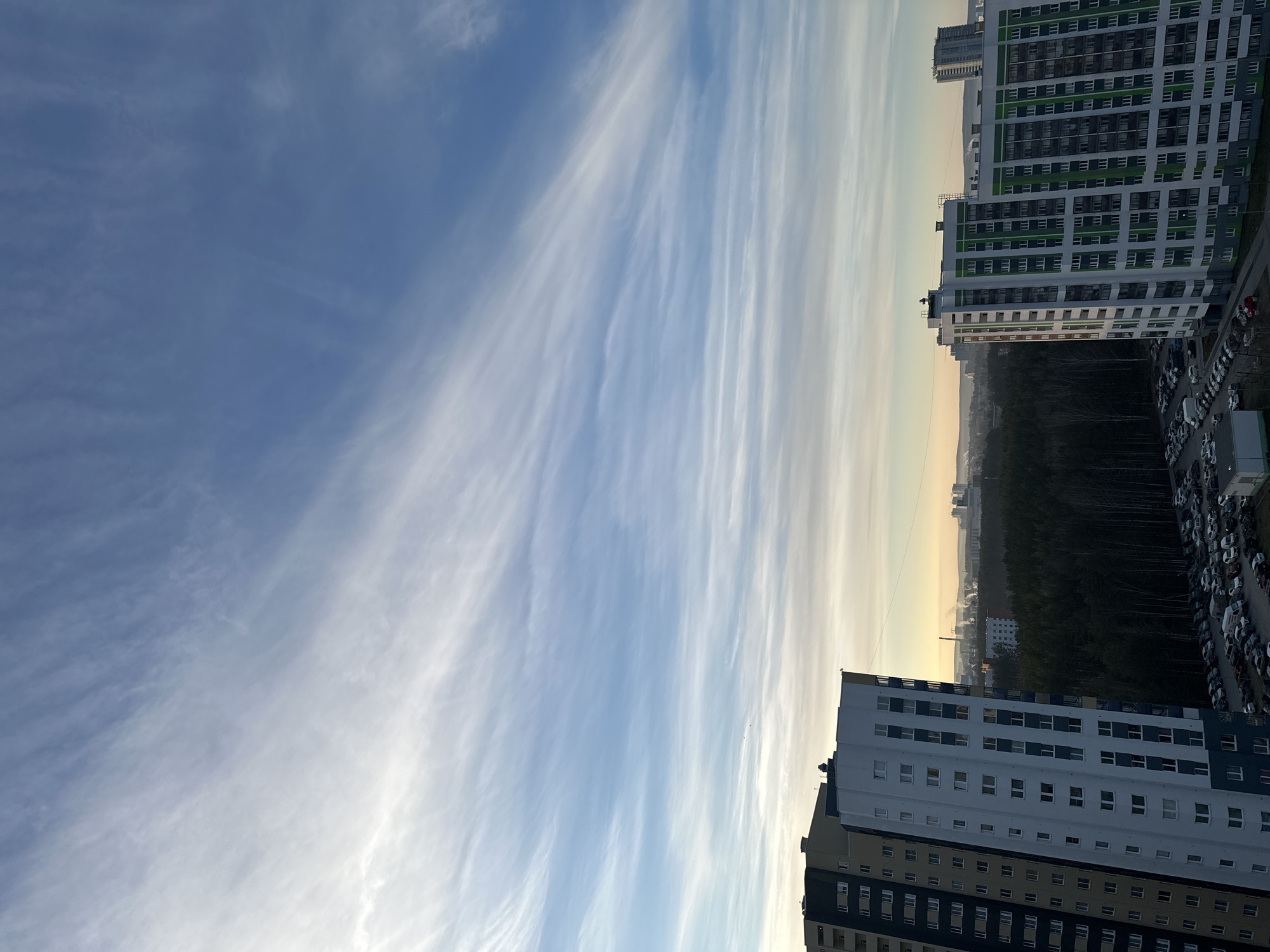 The image shows a view from a high-rise building in Yekaterinburg, Russia. The sky takes up a significant portion of the frame, filled with striated cirrus clouds oriented vertically in the image. Two tall, modern apartment buildings are visible on either side of the image. The building on the right has a white and green color scheme with multiple windows, and a parking lot full of vehicles is visible to its right. The building on the bottom has a white and dark blue color scheme with numerous windows. Between the buildings, a forest line is apparent, with hints of other buildings and possible industrial structures in the distance. The sky transitions to a lighter shade near the horizon, indicating sunrise or sunset.