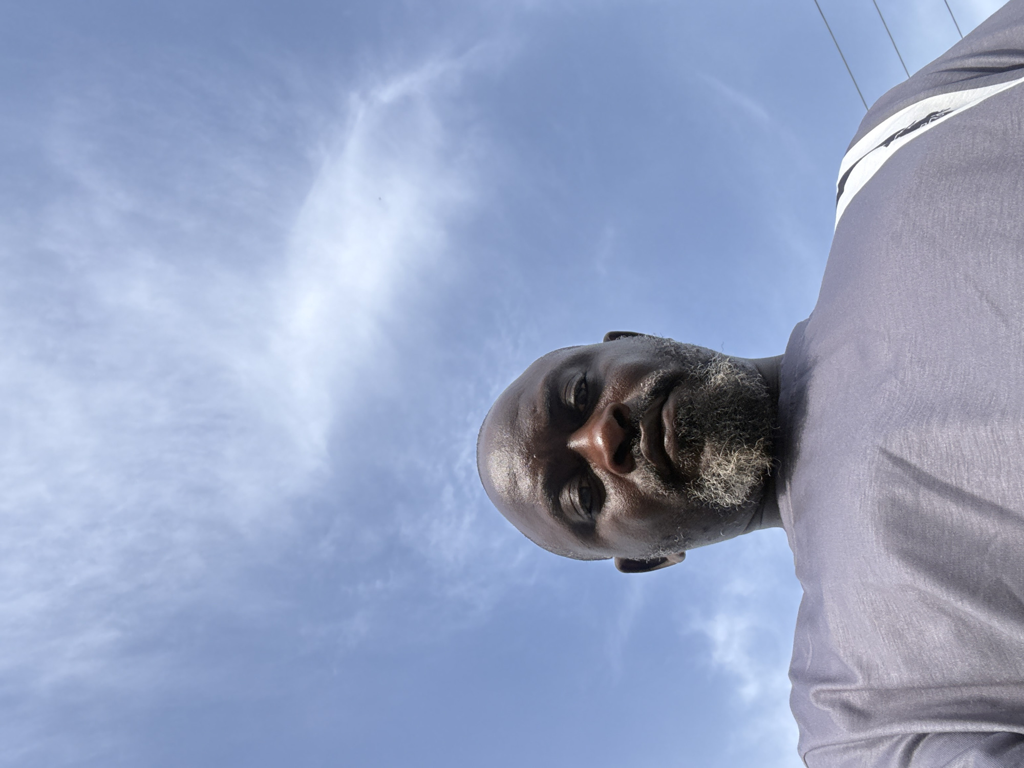 The image captures an individual, presumably male, with dark skin, a shaved head, and a short, salt-and-pepper beard. He is wearing a light grey or lavender t-shirt featuring a white stripe design on the upper right shoulder. The person is positioned in the upper right portion of the frame, photographed from a low-angle perspective looking upwards. His gaze is directed downwards towards the camera, and his face appears somewhat shiny, possibly from sweat or bright light.
The background is a vast, clear blue sky with scattered, wispy white clouds, indicating bright daylight conditions. Faint overhead power lines are visible against the sky in the upper right, suggesting an outdoor, possibly urban or semi-urban environment. There is no specific activity depicted beyond the individual posing for the photo. No visible text is present in the image. The location context provided is Ado, Nigeria.