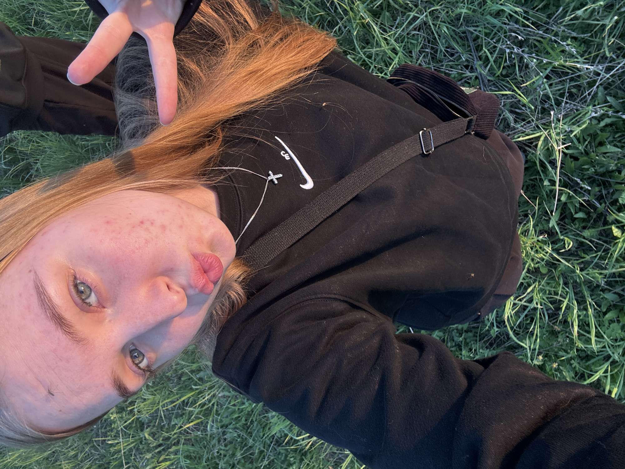 A young person lies on their back in grass, positioned at a slight angle to the camera. They are wearing a black Nike sweatshirt with a silver cross necklace. The individual's hand is raised, making a peace sign. The surrounding environment is characterized by grass and a bag. There is no indication of this image being in Zhigalki, Russia.