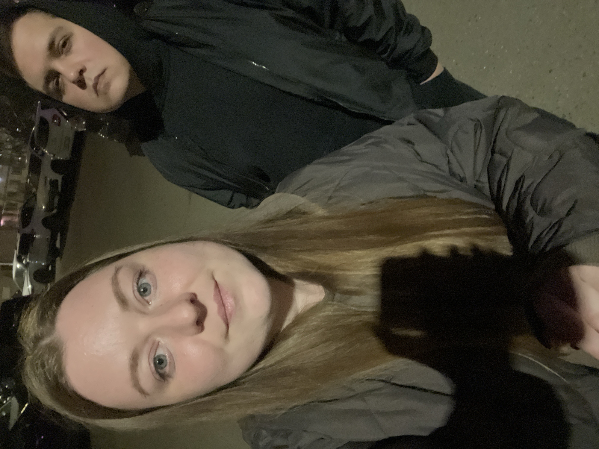 Two people stand closely together, likely taking a selfie. The foreground shows a woman with long blonde hair wearing a grey, quilted jacket. Her face is illuminated, revealing light eyes and a neutral expression. Behind her is a person wearing a black hooded jacket. Their face is partially obscured by the hood and shadows, but their expression appears serious. To the left of the frame, cars are parked along a street, possibly indicating an urban environment. The lighting suggests it is evening or nighttime. This image contains no information to connect the location of Klin, Russia.