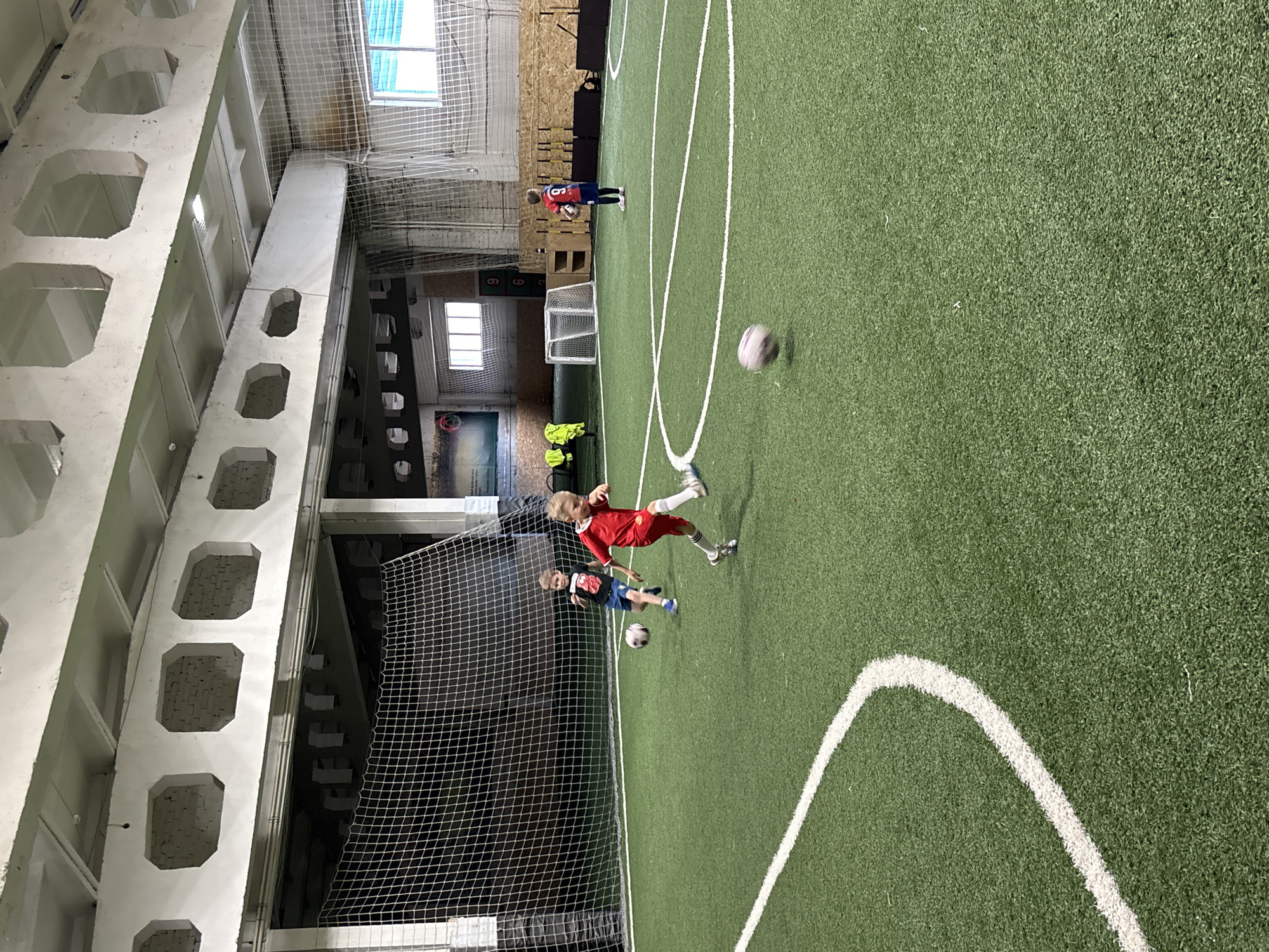 An indoor soccer field in Chaikovsky, Russia, is depicted with three young boys playing. Two boys are actively engaged in a soccer game in the foreground, one in a red jersey kicking a ball, and another in a black t-shirt running. A third boy in a blue and red jersey with the number 9 is standing in the mid-ground holding a soccer ball. The field is artificial turf with white boundary lines. The background shows a netted enclosure, a small soccer goal, and the facility's industrial-style ceiling with exposed beams and lighting.