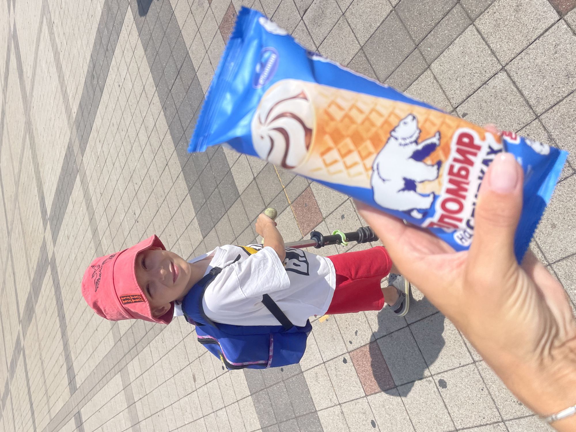 A young boy wearing a red bucket hat, white t-shirt, red shorts, and grey crocs stands on a paved area, holding onto a scooter. He is looking towards the camera with a slight smile. He is also wearing a blue backpack. In the foreground, a hand is holding a blue ice cream wrapper. The ice cream wrapper has an image of a polar bear and text that reads "пломбир" (plombir) and "Maximum". The paved area is made of square grey tiles with darker grey grout lines. The scene appears to be outdoors on a sunny day. Based on the text on the ice cream wrapper, the image likely originates from Russia, and the text "Novorossiysk" appears in a label on the ice cream wrapper.