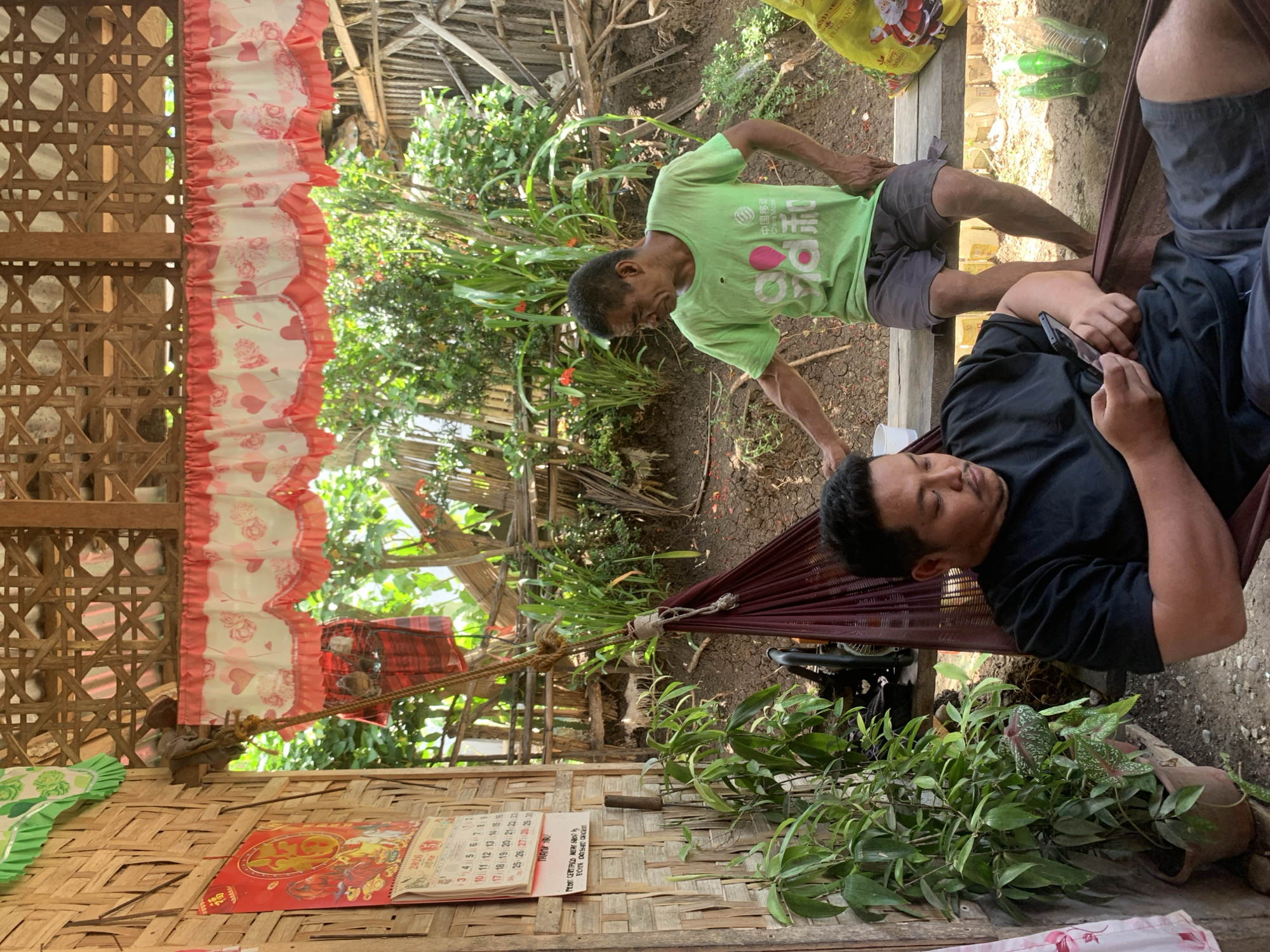In Kiblawan, Philippines, two men are seen in a rustic, open-air dwelling. The man in the foreground, wearing a black t-shirt and grey shorts, is reclining in a burgundy hammock, engrossed in his smartphone. To his right, a second man, in a light green t-shirt and grey shorts, sits on a wooden bench. He is gesturing towards the first man, possibly in conversation. The background reveals lush green foliage and a rustic bamboo wall. A calendar displaying the year 2020 hangs on the wall to the left. The environment suggests a rural or semi-rural setting, possibly a backyard or outdoor common area.