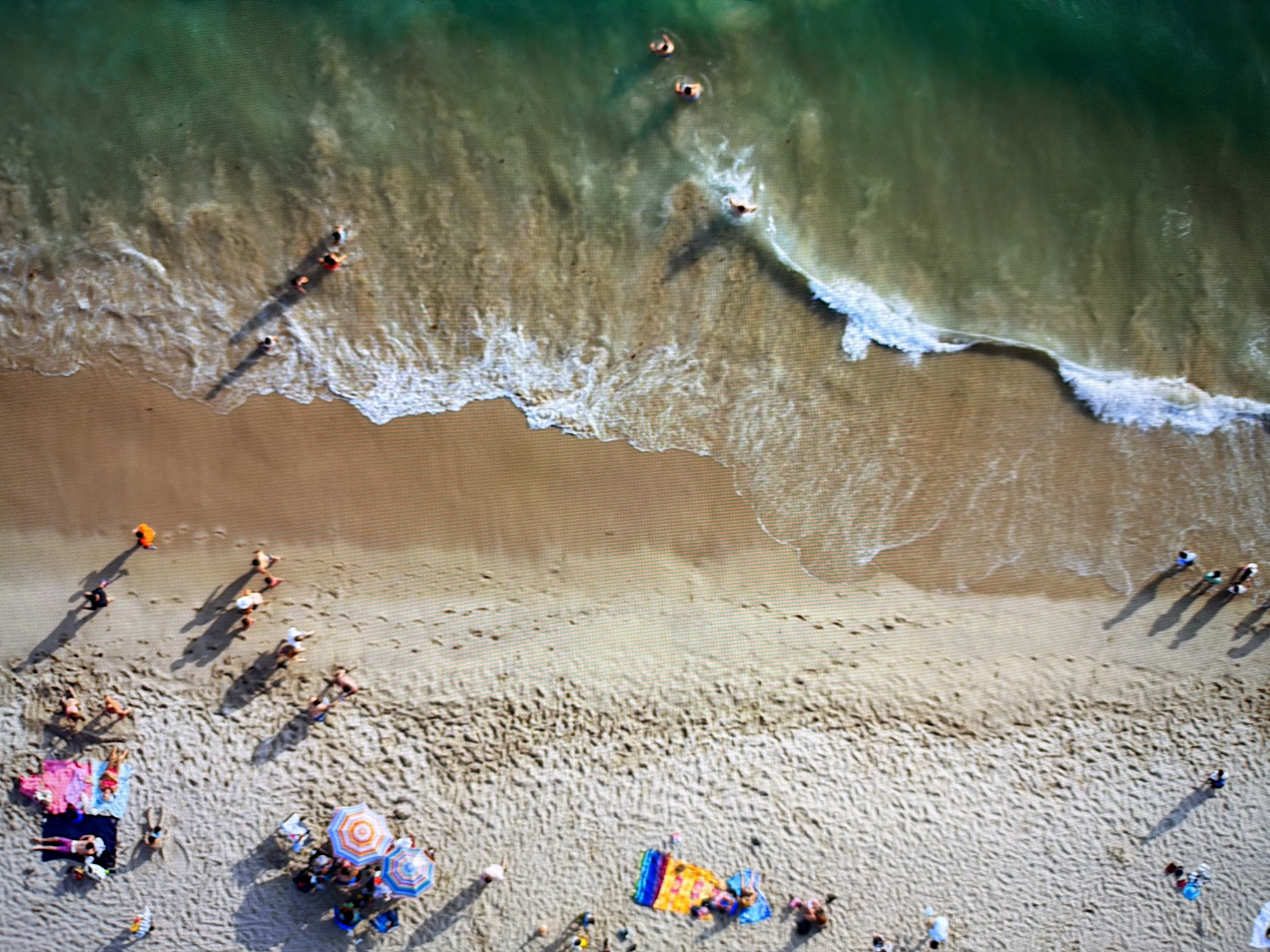 This is an aerial view of a beach scene. On the left side of the image is a sandy beach with several groups of people. Some people are lying on towels, while others are standing or walking. There are also beach umbrellas and a colorful beach mat. On the right side of the image is the ocean with gentle waves lapping at the shore. Several people are visible in the water, swimming or wading. The water is a clear green color, and the sand is a light beige. The lighting suggests it is daytime, likely with bright sunshine, casting shadows on the sand. The scene depicts a typical summer day at the beach with recreational activities. The location is not Kyiv, Ukraine.