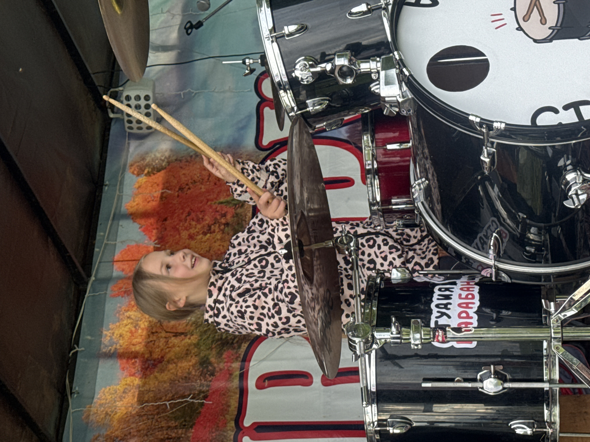 A young female, appearing to be between 8 and 12 years old, with light brown hair, is positioned behind a black and chrome drum set on what appears to be an outdoor stage. She is wearing a pink and black leopard-print hooded tracksuit and holds a wooden drumstick in each hand, raised above the cymbals. Her head is turned towards her right (viewer's left), and she exhibits a slight smile. The drum set includes multiple black tom-toms, a red floor tom partially visible to the left, and a bass drum with a white head featuring a circular port and a stylized cartoon drum graphic. A white sticker with Cyrillic text "СТУДИЯ БАРАБАН" (translating to "DRUM STUDIO") is affixed to one of the black tom-toms. Hi-hat cymbals are visible to the left, and a larger crash or ride cymbal is positioned to the right. The background consists of a stage backdrop depicting an autumn landscape with orange and yellow trees against a light blue sky, and a large red and white letter "P" graphic is partially visible on the left side of the backdrop. Above the backdrop, a dark, fabric-covered structure, likely a stage canopy, is visible, along with white string lights and a white, gridded stage light fixture. A microphone and stand are partially visible on the right side of the frame. The scene is located in Большая Кивара, Russia.