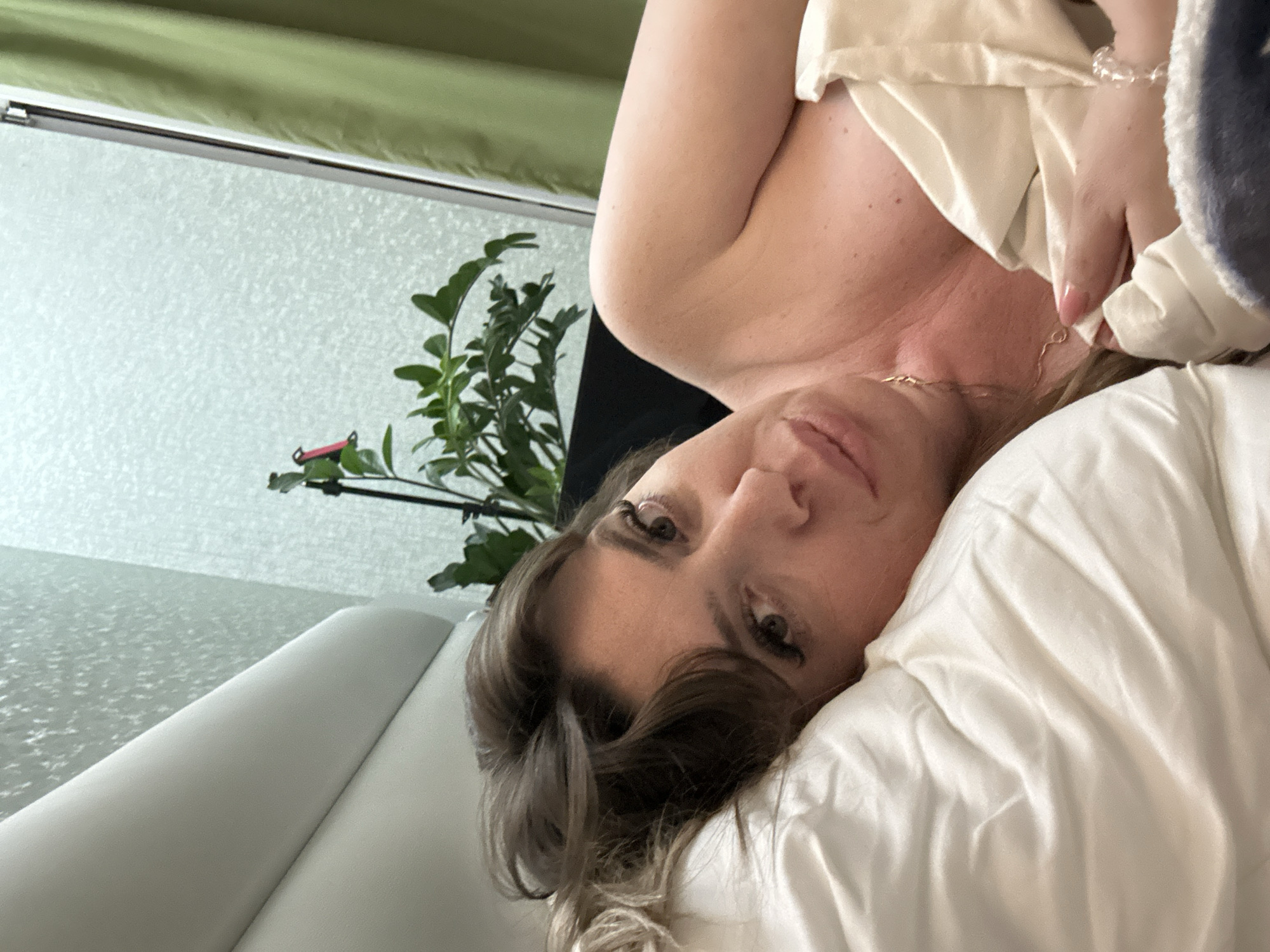 A fair-skinned woman with brown hair lies on a white pillow and is partially covered by a white sheet or garment. She is wearing a necklace. The background includes a white wall, a green curtain or valance, and a green plant. The location cannot be determined from this image.