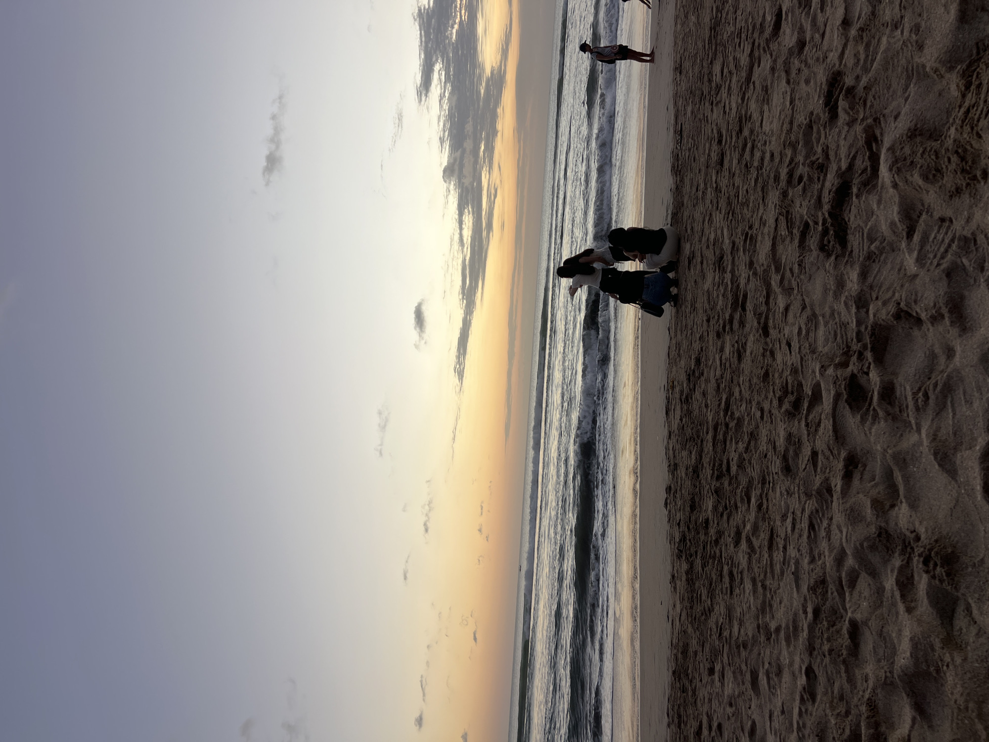 The image depicts a tranquil scene at a beach in Kuta, Indonesia, during what appears to be sunset. The sky transitions from a soft blue at the top to warm hues of orange and yellow near the horizon, with scattered clouds. The ocean displays gentle waves rolling towards the shore, creating a foamy white line where they meet the sand. In the foreground, the textured sand is visible, with footprints and subtle ripples from the receding tide.
Several individuals are present on the beach. In the middle ground, a group of four people are gathered. Two individuals are crouching, while another two stand behind them. One of the standing figures appears to be pointing towards the ocean. To the right, a lone male figure stands facing the sea, and further to the right, another person is partially visible near the water's edge. The overall atmosphere is serene and suggests a moment of shared experience or quiet observation of the natural scenery.