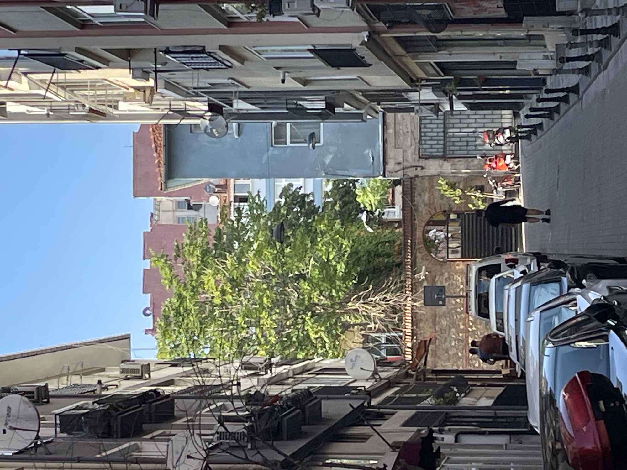 The image shows a narrow street in Kadıköy, Turkey, lined with multi-story buildings. The street is paved and there are parked cars on the left and right sides. Several buildings on the left have satellite dishes mounted on their facades. On the right, a man in a black shirt and shorts is walking away from the camera. Further down the street, two red motorbikes are parked. The street leads to an arched entranceway with a stone wall and a large tree behind it. In the background, additional buildings are visible, including one painted light blue. The sky is clear and blue.