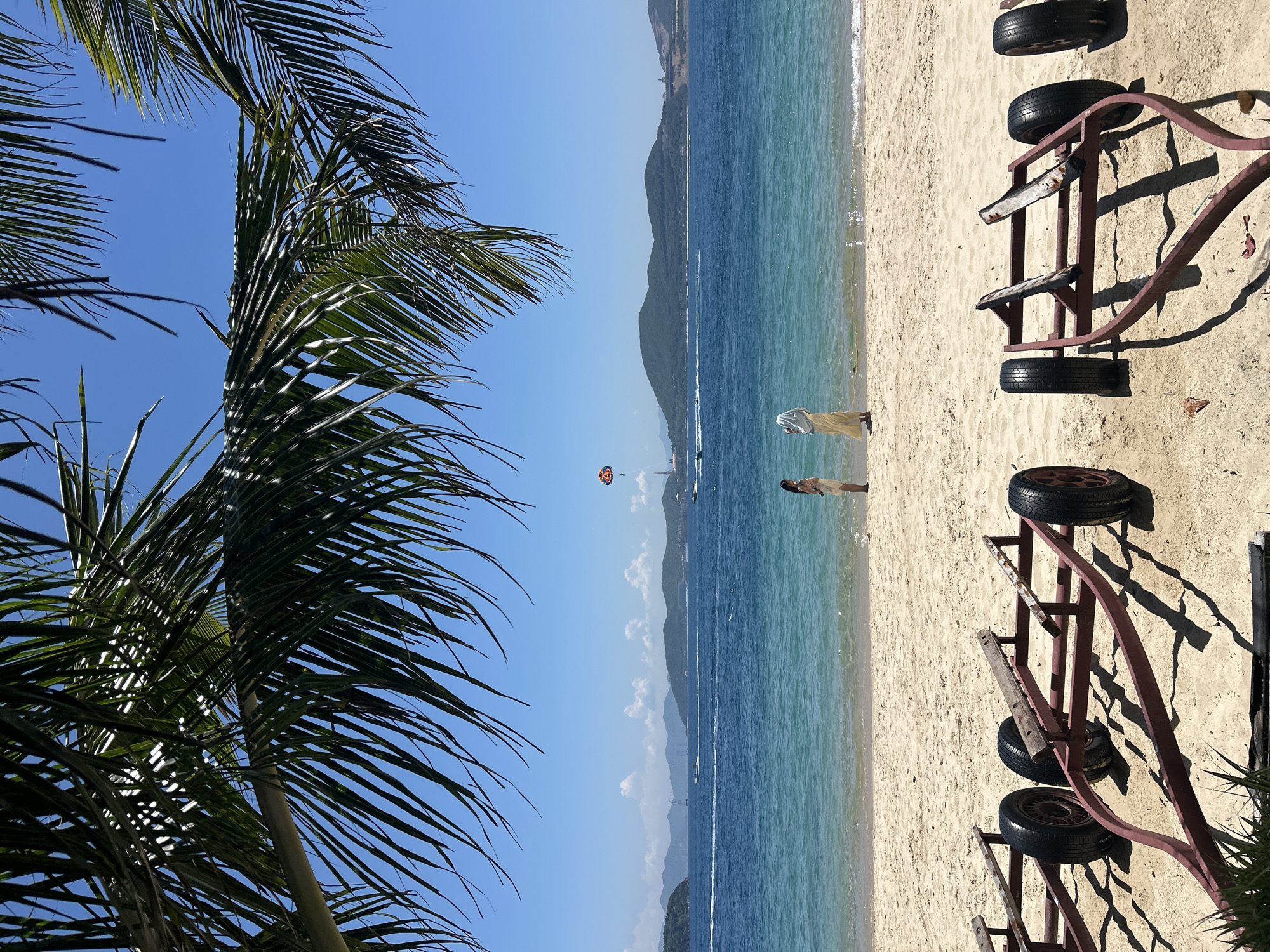 This is a low-angle, full shot of a beach scene in Nha Trang Ward, Vietnam. In the foreground, two weathered, red boat trailers with black tires are parked on the sand. The beach stretches to the ocean, where gentle waves lap the shore. Two individuals stand on the beach, facing the water. One person, wearing a light-colored dress, stands to the left, and another, in a yellow robe and white headscarf, stands to the right. In the distance, the blue ocean meets a hazy horizon with green, mountainous islands. A bright orange parachute is visible in the sky above the water, with a boat or watercraft tethered to it. Several other boats are on the water, creating wakes. Palm fronds from a tree in the upper left frame cast shadows across the image. The sky is a clear, bright blue with a few scattered white clouds.