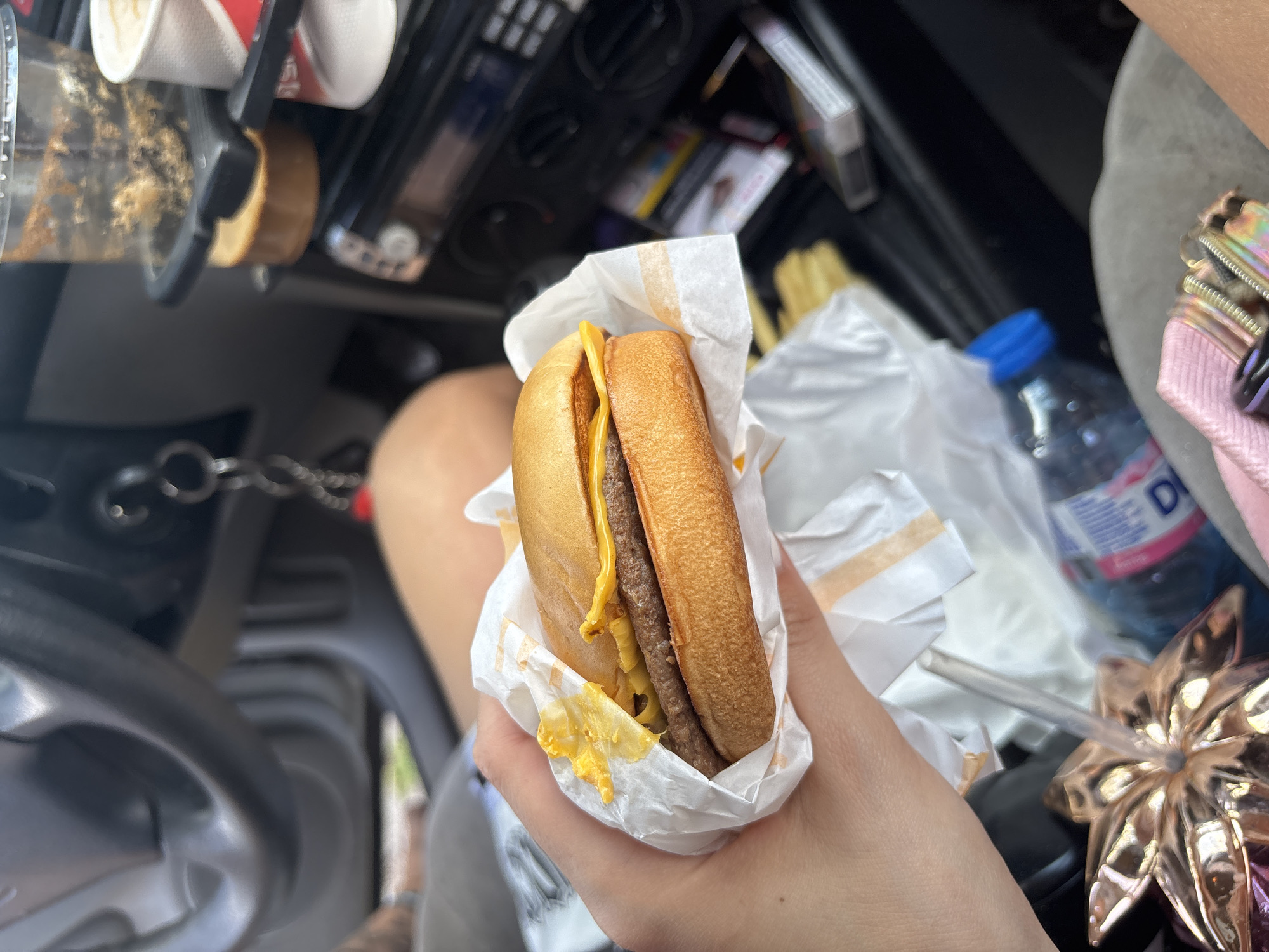 A close-up, eye-level shot captures a person holding a cheeseburger in a car, indicating an on-the-go meal. The focal point is a hand grasping a single-patty cheeseburger wrapped in white paper, revealing melted yellow American cheese and a toasted bun. Some cheese has dripped onto the wrapper.
The setting is the interior of a vehicle, likely a car, with the person appearing to be in the passenger seat. In the blurred background, several items are visible. To the left, a transparent cup with a brown residue (possibly from a coffee or soda) sits in a cup holder near the dashboard. Further back, a blurry view of French fries in a white paper container can be seen. To the right, a clear plastic water bottle with a blue cap is present, along with a decorative, golden, flower-shaped cup or lid with a straw. A pink textured object, possibly a bag or purse, is also partly visible. The car's dashboard, including various controls and the top of a steering wheel (left-hand drive), is partially obscured in the upper left.
The lighting suggests it is daytime. No specific weather conditions are discernible. There is no clearly readable text, except for a partial "D" on the water bottle label.