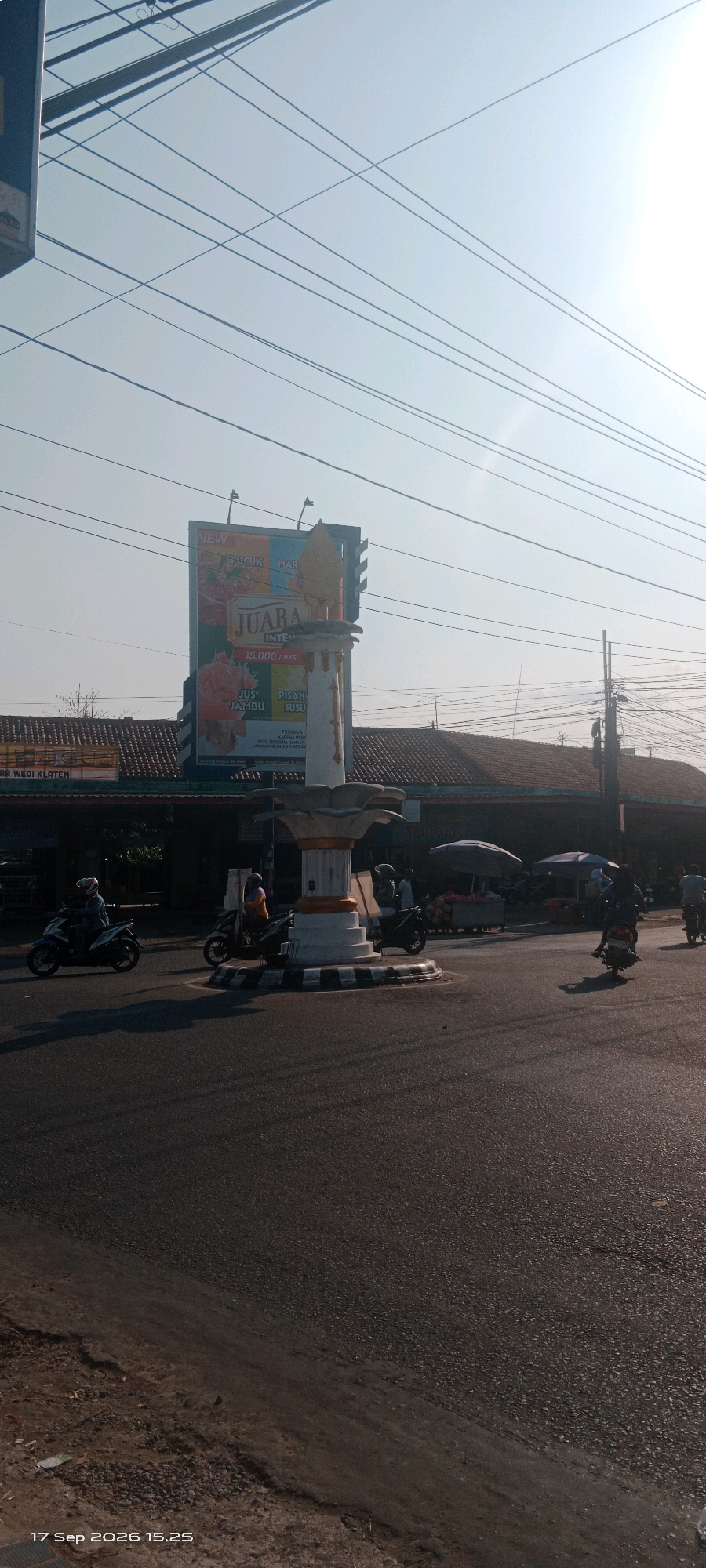 The image shows a roundabout in Wedi, Indonesia, with a monument at its center. Several motorbikes are visible on the road, with riders wearing helmets. A billboard with the word "JUARA" is displayed in the background, along with advertisements for "JU-JAMBU" and "PISAN SUSU". A building with a tiled roof is behind the billboard. Overhead, power lines crisscross the sky. The scene is illuminated by sunlight, casting shadows on the road. A timestamp at the bottom reads "17 Sep 2026 15.25".