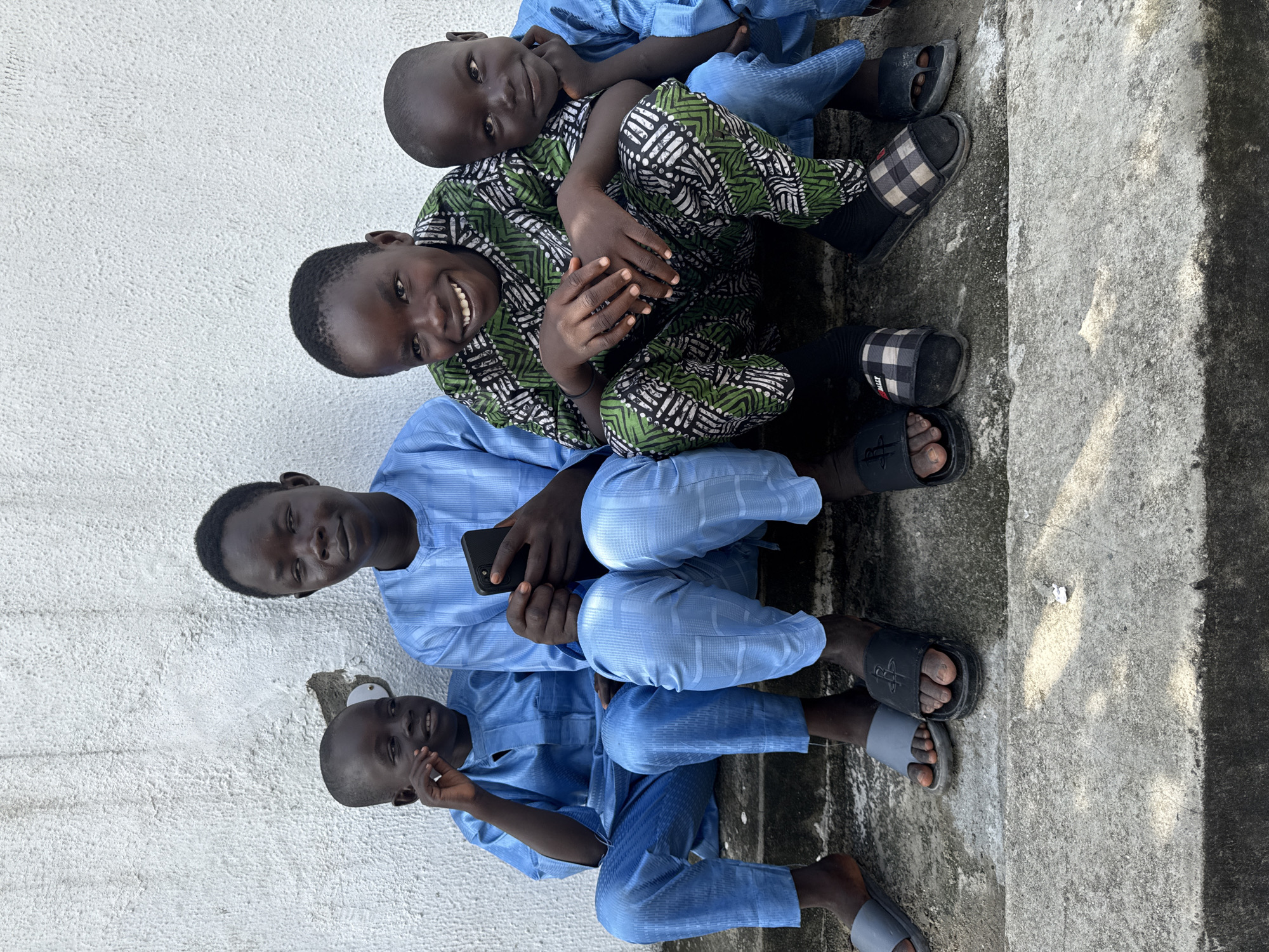 Four young males are sitting on an outdoor concrete surface in Ado, Nigeria. The backdrop includes a white wall to the left. A taller, older male dressed in a light blue patterned shirt and pants sits at the center, holding a dark-colored smartphone. Beneath him and leaning against the wall, another young boy wearing a matching light blue outfit is seated. Above the taller male are two younger boys; the one closest to him is wearing a green and black patterned shirt and dark pants. The boys are all smiling. All four boys are wearing sandals or slippers. The concrete surface has some shadowing and stains.