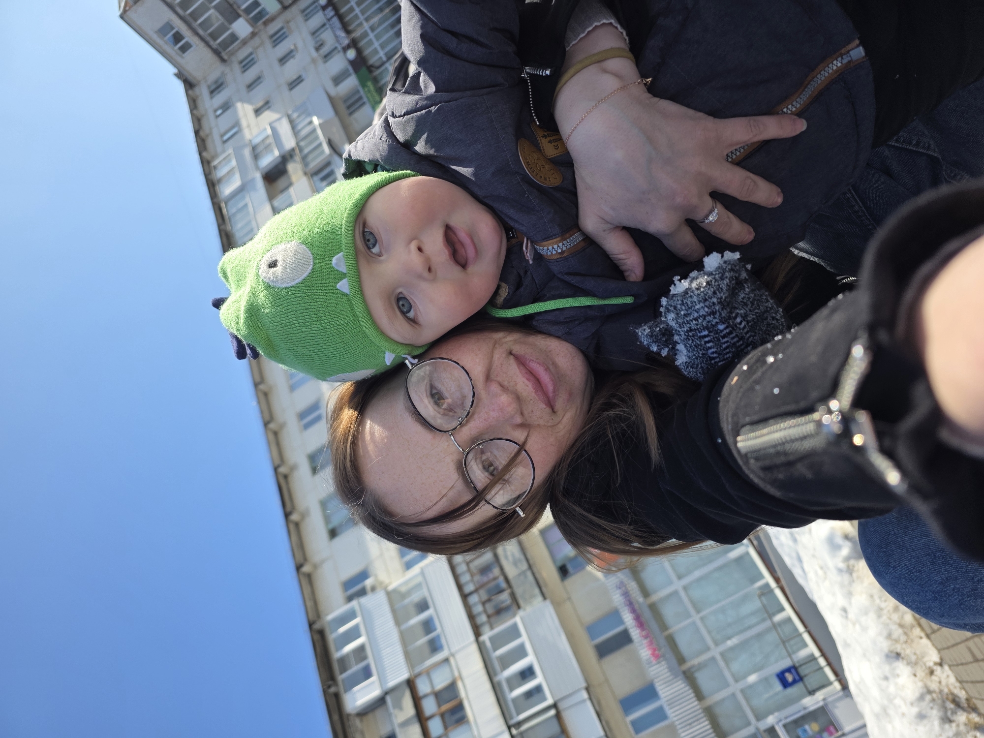 The image captures an adult female and an infant male posing for a selfie outdoors in Chaikovsky, Russia, under a clear blue sky. The female, with brown hair and eyeglasses, is positioned on the left, looking directly at the camera with a slight smile. She is wearing a dark-colored jacket with visible zippers and reflective elements, blue jeans, a silver-colored ring on her left ring finger, and a thin gold-colored chain bracelet and a wider tan band on her right wrist. Her left arm is wrapped around the infant. The infant, positioned to her right, wears a green knitted hat adorned with a white eye and small white teeth-like features, along with blue spikes on top, resembling a monster or dinosaur. He has blue eyes and an open mouth, also looking towards the camera, and is dressed in a dark grey winter jacket with a brown patch on the chest and a green drawstring.
The background reveals a multi-story, light-colored residential building typical of urban Russian architecture, featuring numerous windows and enclosed balconies. On the ground floor of this building, a storefront with large glass windows and a pink "Wildberries" sign is visible, along with a blue box, possibly a mailbox or ATM, near its entrance. Patches of snow and bare ground are present in the immediate foreground, indicating a cold weather season such as late winter or early spring.