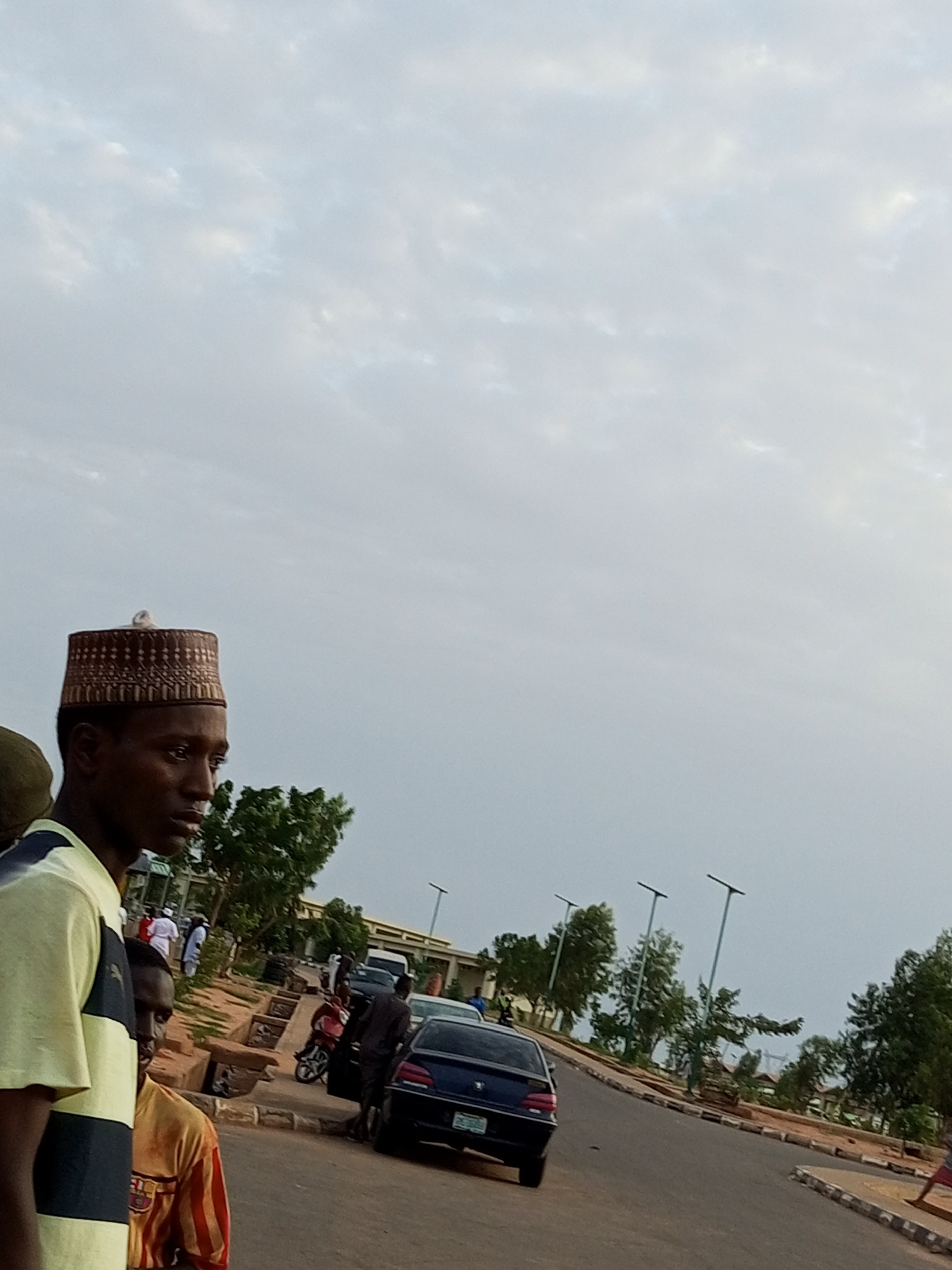 The image depicts a street scene in Gombe, Nigeria. In the foreground, a young man wearing a patterned cap and a striped t-shirt is shown in profile, looking to the right. Behind him, another person in a striped shirt is partially visible. The background shows a road curving to the right, with several parked vehicles including a dark sedan and a motorcycle. People, some dressed in white traditional attire, are visible near buildings in the distance. Trees line the side of the road, and streetlights are present. The sky is overcast.