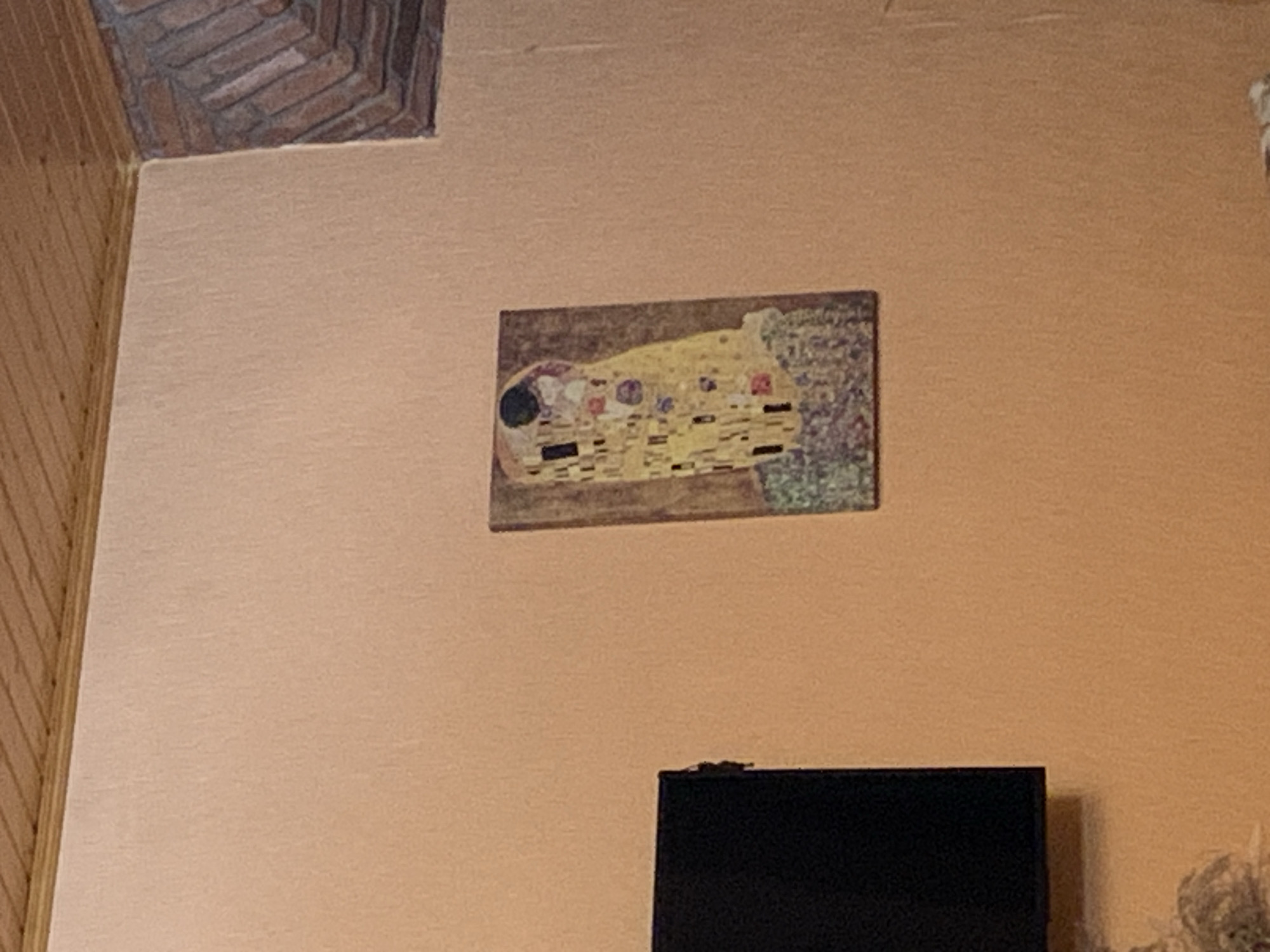 A framed painting, recognizable as Gustav Klimt's "The Kiss," is displayed on a peachy-orange wall. The painting is oriented vertically and features two figures embracing, adorned with intricate patterns and gold leaf. To the left of the painting, a portion of a black rectangular object, likely a television, is visible. The wall itself has a textured surface. Above the wall, a wooden ceiling with horizontal planks is present. In the upper right corner, a decorative architectural detail made of red bricks is integrated into the wall. The overall scene suggests an indoor setting.