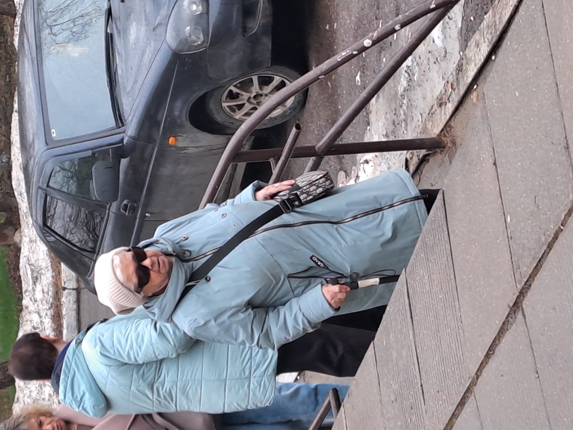 The photograph captures a street scene in Elektrostal, Russia. An adult female wearing a light blue quilted coat, sunglasses, and a white knit cap is situated on a staircase next to a black car. The woman has a purse slung across her body and appears to be holding an object in her right hand. A man with dark hair is partially visible to her left, and the legs of another person are visible below him. The scene includes metal railings and concrete stairs. The background shows a portion of the dark car and other indiscernible elements of the street.