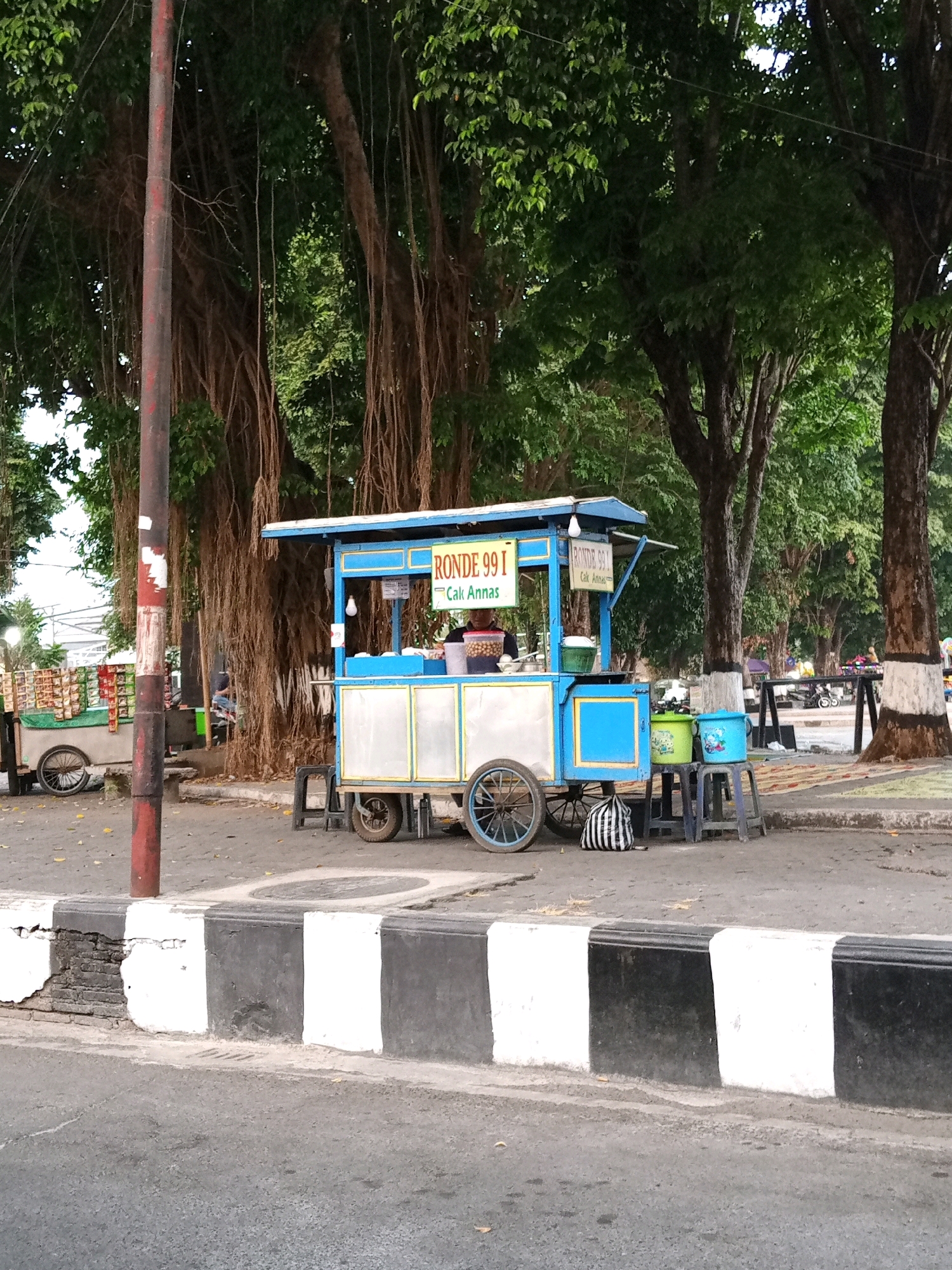 A street scene in Sukoharjo, Indonesia, features a blue and white food cart with the text "RONDE 99 I Cak Annas" prominently displayed. The cart is positioned on a paved area adjacent to a sidewalk marked with alternating black and white painted blocks. Behind the cart, large trees with hanging roots create a shaded canopy. To the left of the main cart, another smaller cart is visible, laden with various food items. A person is partially visible behind the main cart, appearing to be attending to the food. In the background, more trees line the street, and glimpses of other street vendors and passing vehicles can be seen. The ground is a mixture of pavement and dirt, with scattered fallen leaves.