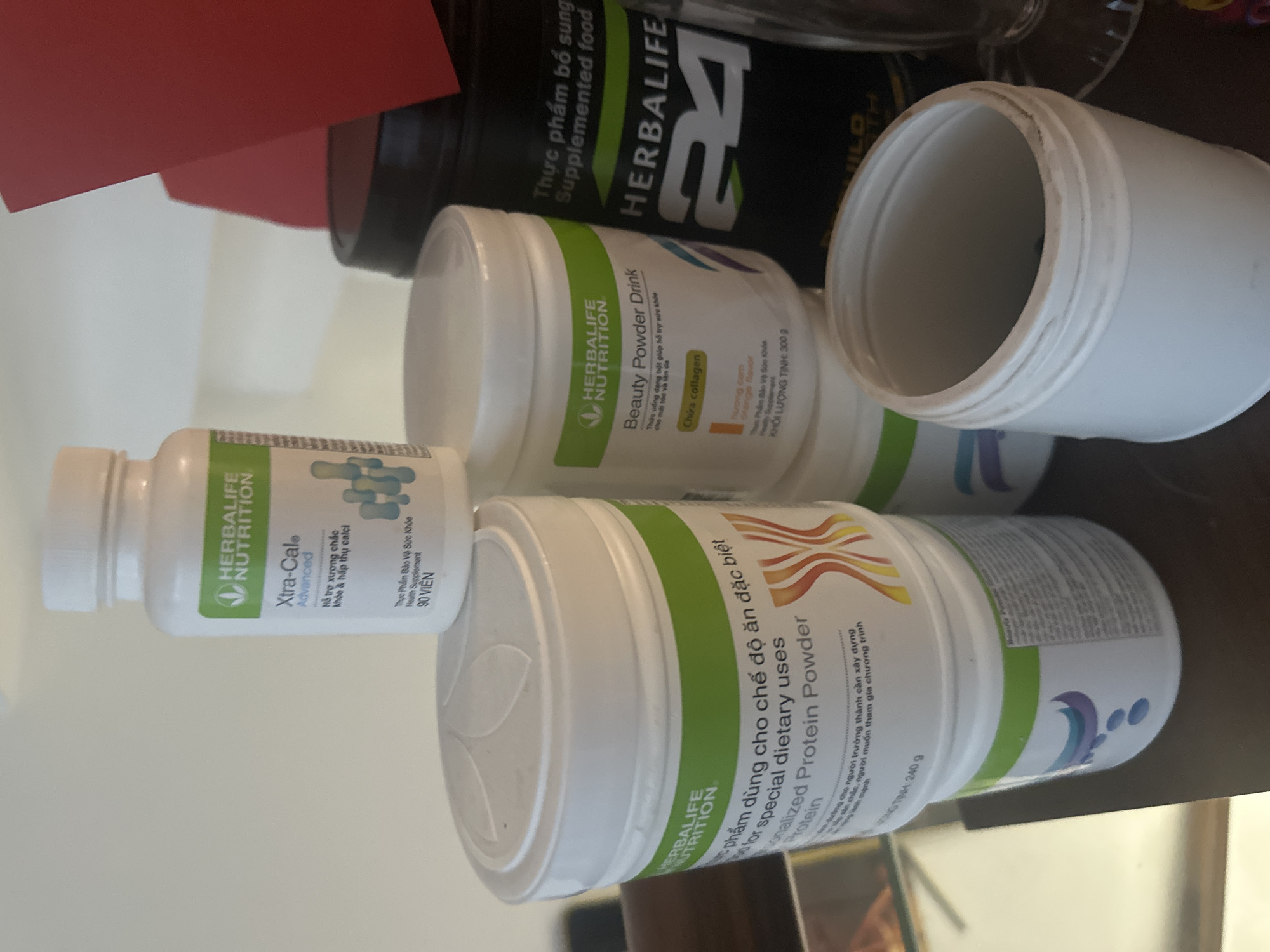 The image shows several containers of Herbalife Nutrition products. There is a black container of "Supplemented food" and a white container of "Beauty Powder Drink," both labeled "HERBALIFE NUTRITION." There is a small, white bottle labeled "Xtra-Cal Advanced" containing 90 tablets. Also present is a white container labeled "personalized protein powder." An empty, open white container sits on top of the others. The location is suggested to be Bac Nha Trang Ward, Vietnam based on the Vietnamese text on the containers.