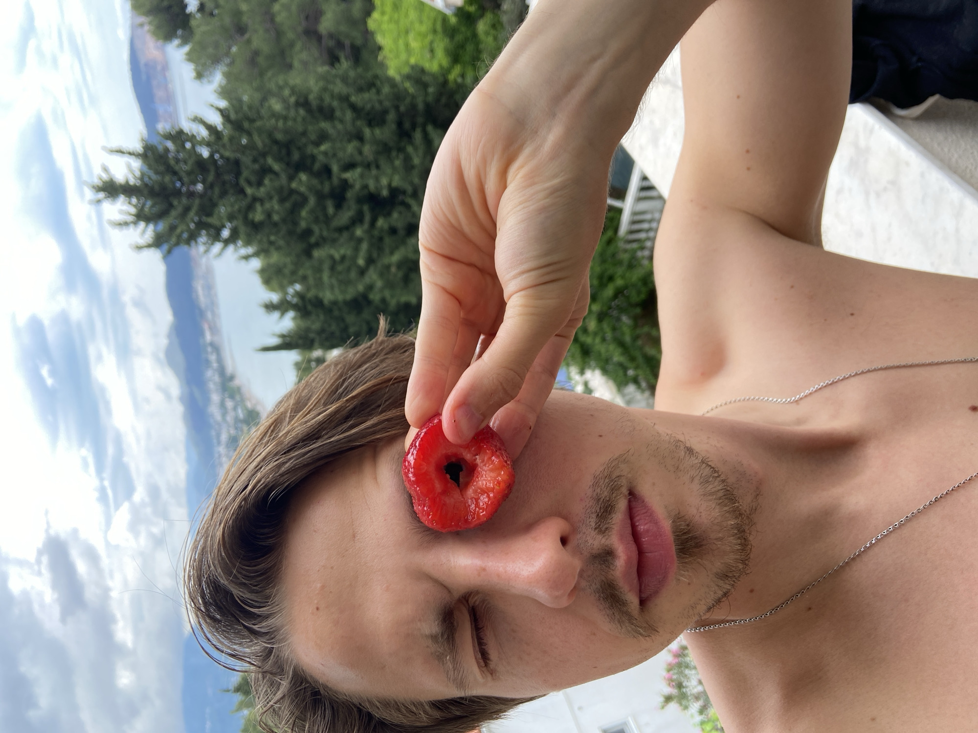 A man with light brown hair and a stubbly beard is holding a cored strawberry in front of his closed left eye. He is shirtless and wearing a silver chain necklace. The background shows greenery, the sky, and a body of water with a city visible in the distance. This is a view of Šušanj, Montenegro.