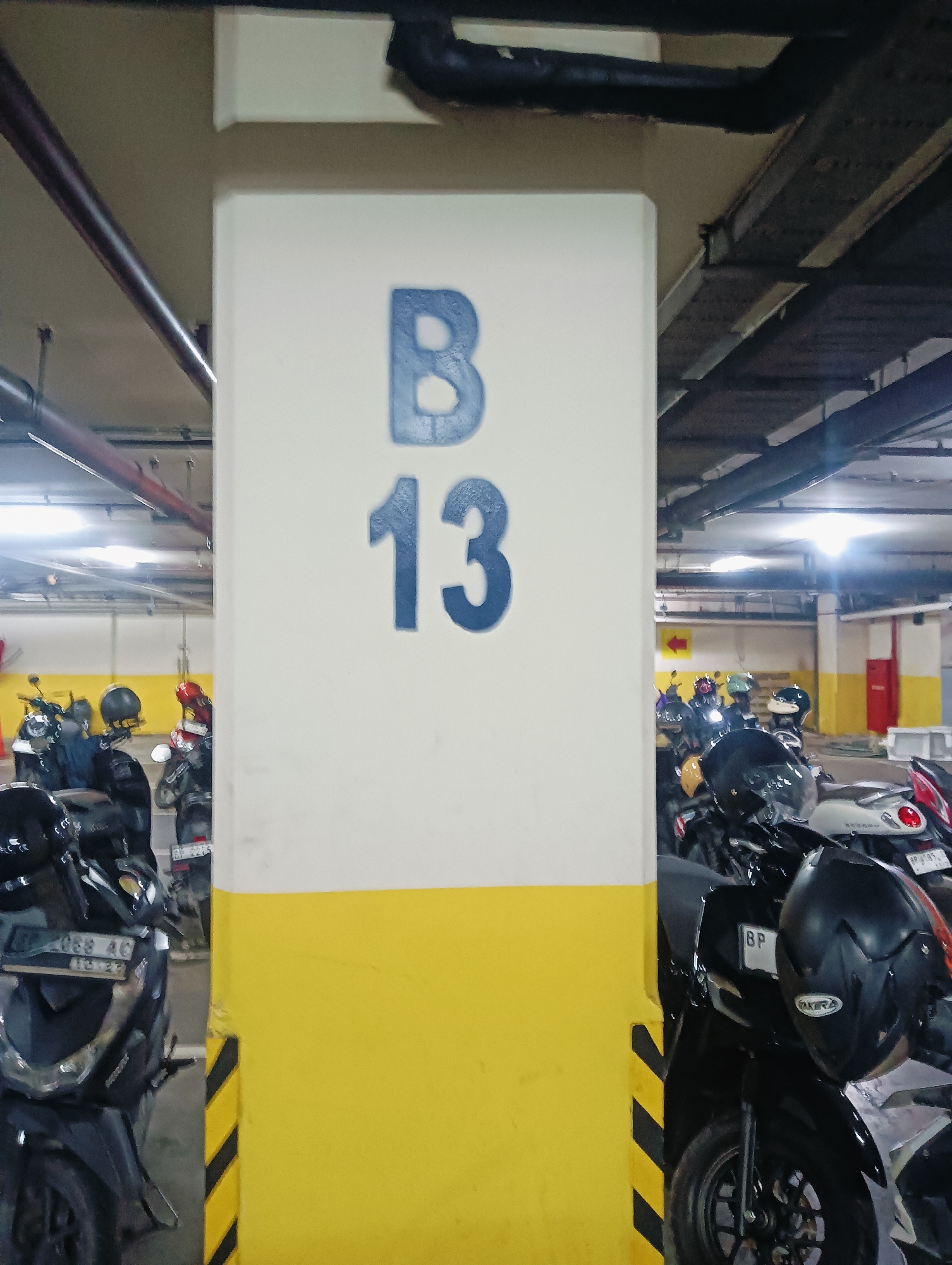 The image captures a parking area, likely indoors, possibly a multi-story car park given the visible piping and lighting fixtures. Dominating the center of the frame is a thick concrete pillar painted with a large blue letter "B" and the number "13", indicating a parking zone. The pillar is bisected horizontally, with the top half painted white and the bottom half painted yellow with diagonal black stripes. To the left and right of the pillar, numerous parked motorcycles are visible. Several motorcycles are black, some with visible license plates. Helmets of various colors, including black, green, and red, are placed on the seats or handlebars of the motorcycles. The lighting in the background suggests fluorescent or LED lamps, common in such indoor parking facilities. The overall impression is one of an organized, albeit somewhat dimly lit, parking space for two-wheeled vehicles. It is highly probable this is in Batam, Indonesia, a city known for its extensive use of motorcycles as a primary mode of transportation and its numerous commercial and residential complexes that require such parking facilities.