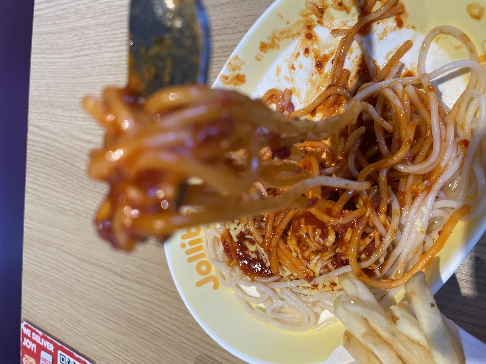 A close-up shot shows a fork lifting a portion of Jollibee spaghetti. The spaghetti is covered in a sweet and savory red sauce with shredded cheese. The plate, with the Jollibee logo visible, contains more spaghetti and a serving of french fries. The setting appears to be indoors, with a light-colored wooden table. The image is well-lit, suggesting it was taken during the day. The focus is on the food, creating an appetizing visual. The text "Jollibee" is visible on the plate. Another piece of text visible at the bottom left corner says "WE DELIVER JOY!". The location is specified as Davao City, Philippines.