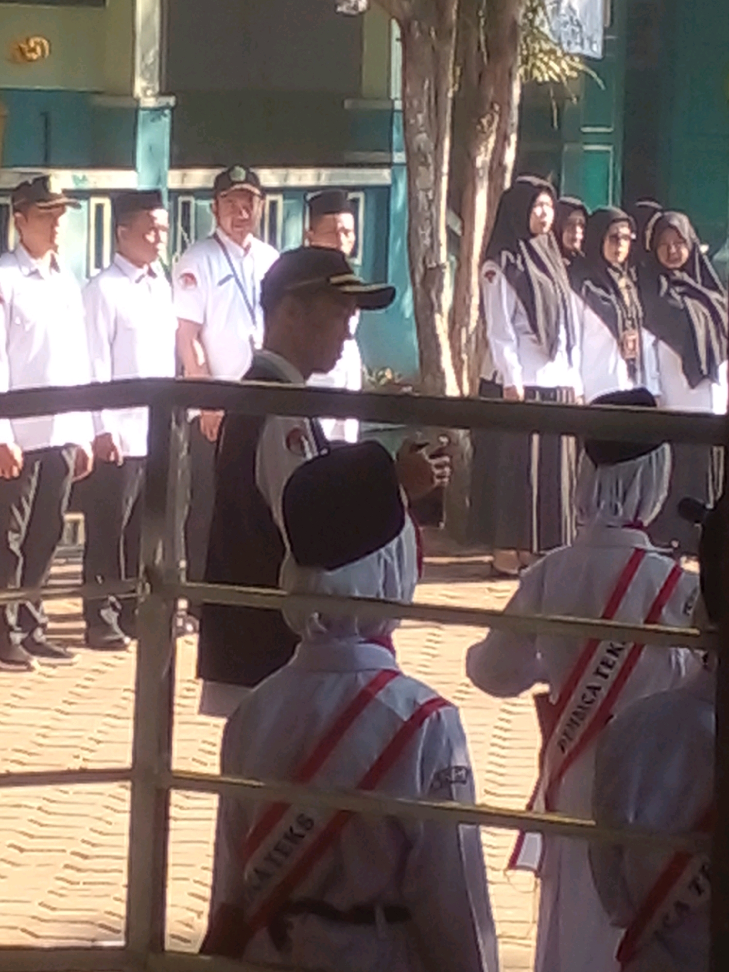 The image shows a group of people standing in what appears to be an outdoor school setting in Banyuputih, Indonesia. Several individuals are wearing white shirts and dark pants. Some of these individuals are wearing caps. A group of women in the background are wearing hijabs. Some of the individuals in the foreground are wearing white uniforms with red stripes, and some have sashes across their chests with text that is partially visible. The background features a building with light blue and green accents, and trees. The ground is paved with patterned bricks. The scene is viewed through a railing.