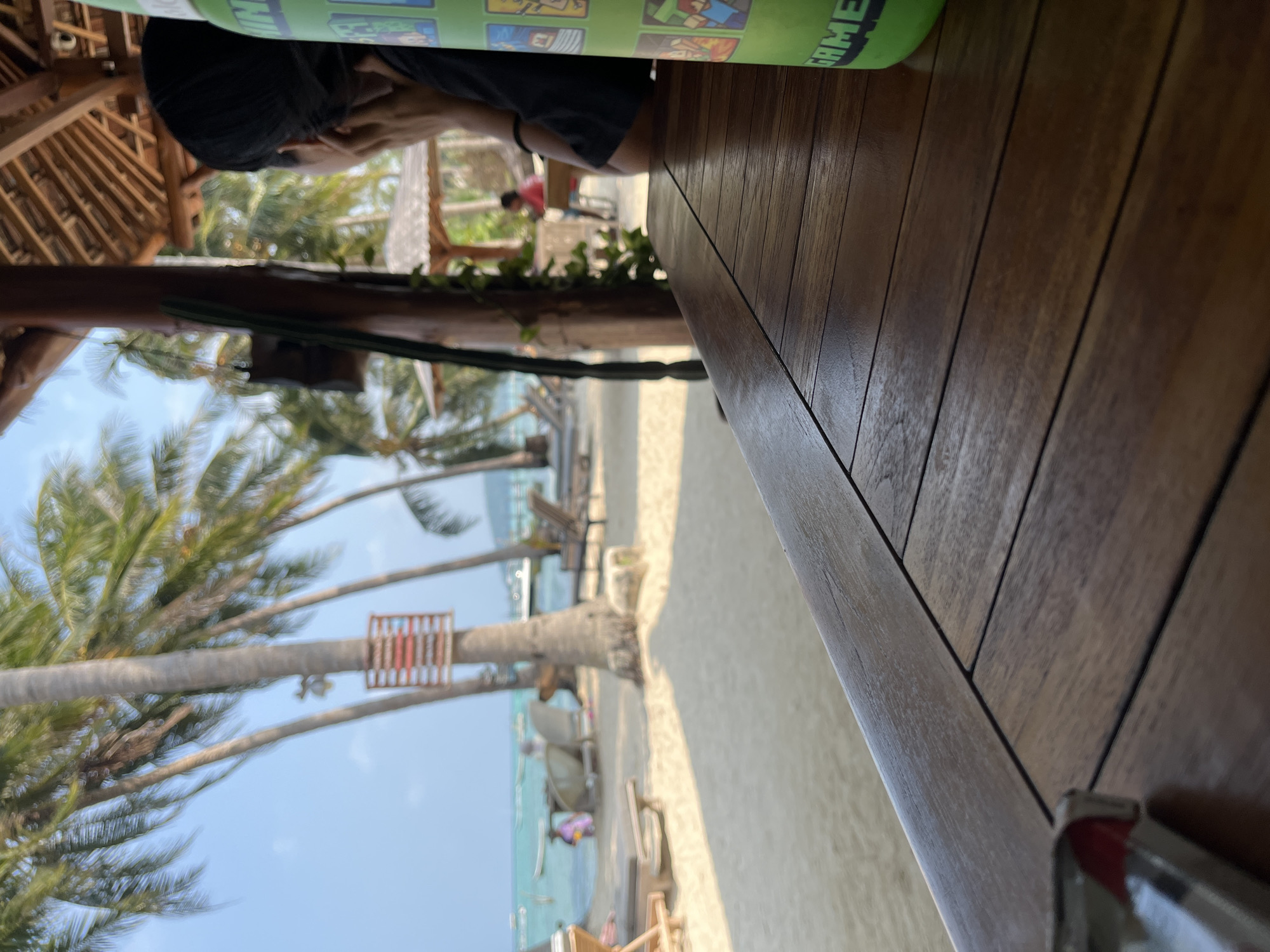 The image depicts a beachside scene with a wooden table in the foreground, suggesting a cafe or restaurant setting. Beyond the table, a sandy beach stretches towards the ocean, dotted with palm trees, lounge chairs, and a few people. In the background, the ocean is visible with small boats. The sky is clear and blue, indicating fair weather. On the right side of the foreground, a green cylindrical object with colorful graphics, possibly a water bottle or a container, is present. The perspective is from a seated position, looking outwards. The overall impression is a relaxed, tropical environment, likely in Legonipah, Indonesia, given the presence of palm trees and the beach setting.