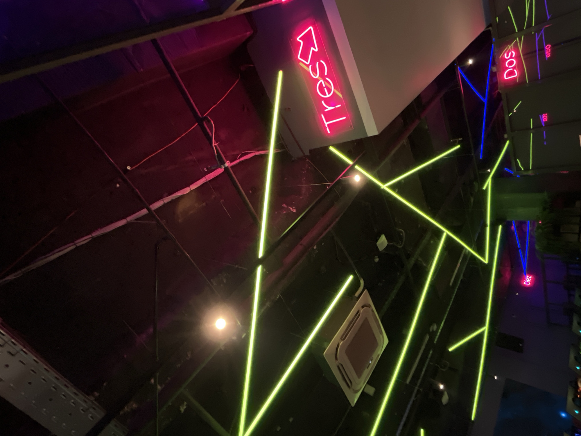 The image shows a ceiling with neon lights in a building. "Tres" is written in red neon with an arrow pointing upwards, and there are green neon lines arranged in angular patterns. An air conditioning unit is visible on the ceiling. Other neon signs say "Dos" and "Diez." The building is in Kazan, Russia.