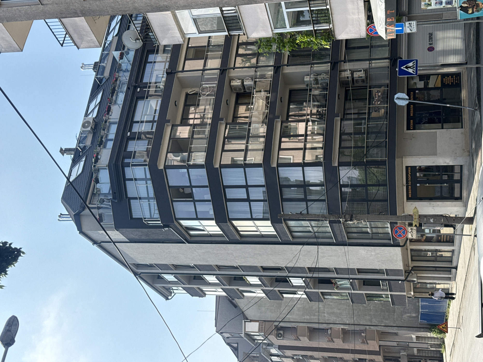 The image captures a sunny street scene in Varna, Bulgaria, dominated by a modern, multi-story building. This prominent structure features a contemporary design with a dark gray facade, large reflective glass windows, and light gray architectural elements. Balconies with glass railings are visible on the upper floors, some adorned with potted plants, and multiple air conditioning units are mounted on the exterior. At least one satellite dish is also visible on an upper balcony.
At street level, the building houses commercial establishments. A shop sign prominently displays "МАРКЕТ" (Market). Another shop window advertises "КУХНИ МЕБЕЛИ БРАТИ" (Kitchens Furniture Brati), with the additional text "ПО ПОРЪЧКА" (By Order) below it. Further down, a metal shutter or sign has the text "НЕ СПИРАЙ!" (Do Not Stop!).
The street area features various traffic signs: a blue square with a white pedestrian crossing symbol, a red circular sign with a diagonal line (No Parking/Stopping), and a blue square with a downward white arrow which is typically a "No Entry" or "Entry Forbidden" sign. Overhead utility wires crisscross the clear blue sky. A lone person, dressed in a light-colored top and dark trousers, is visible walking on the sidewalk. An older building with balconies is partially visible in the background, contrasting with the modern primary structure. The bright sunlight suggests it is daytime with clear weather.