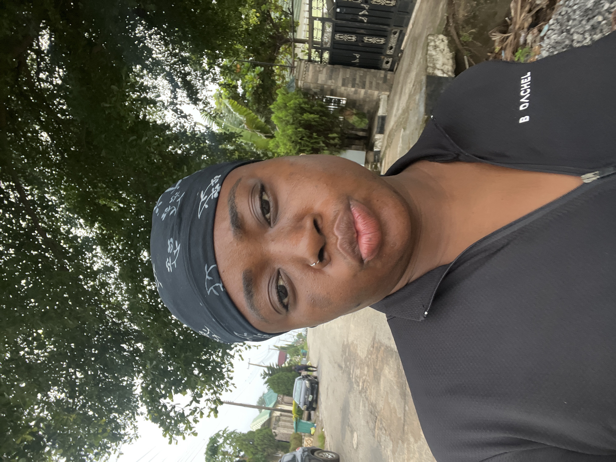 A close-up, slightly rotated, portrait photograph depicts a person with dark skin wearing a black bandana with a light-colored pattern and a black athletic shirt with the name "B DACHEL" printed on the right side of the chest. The person has a nose ring. The background shows a street in Abuja, Nigeria, featuring vegetation, houses, a gate, parked cars, and pedestrians in the distance. The day is overcast.