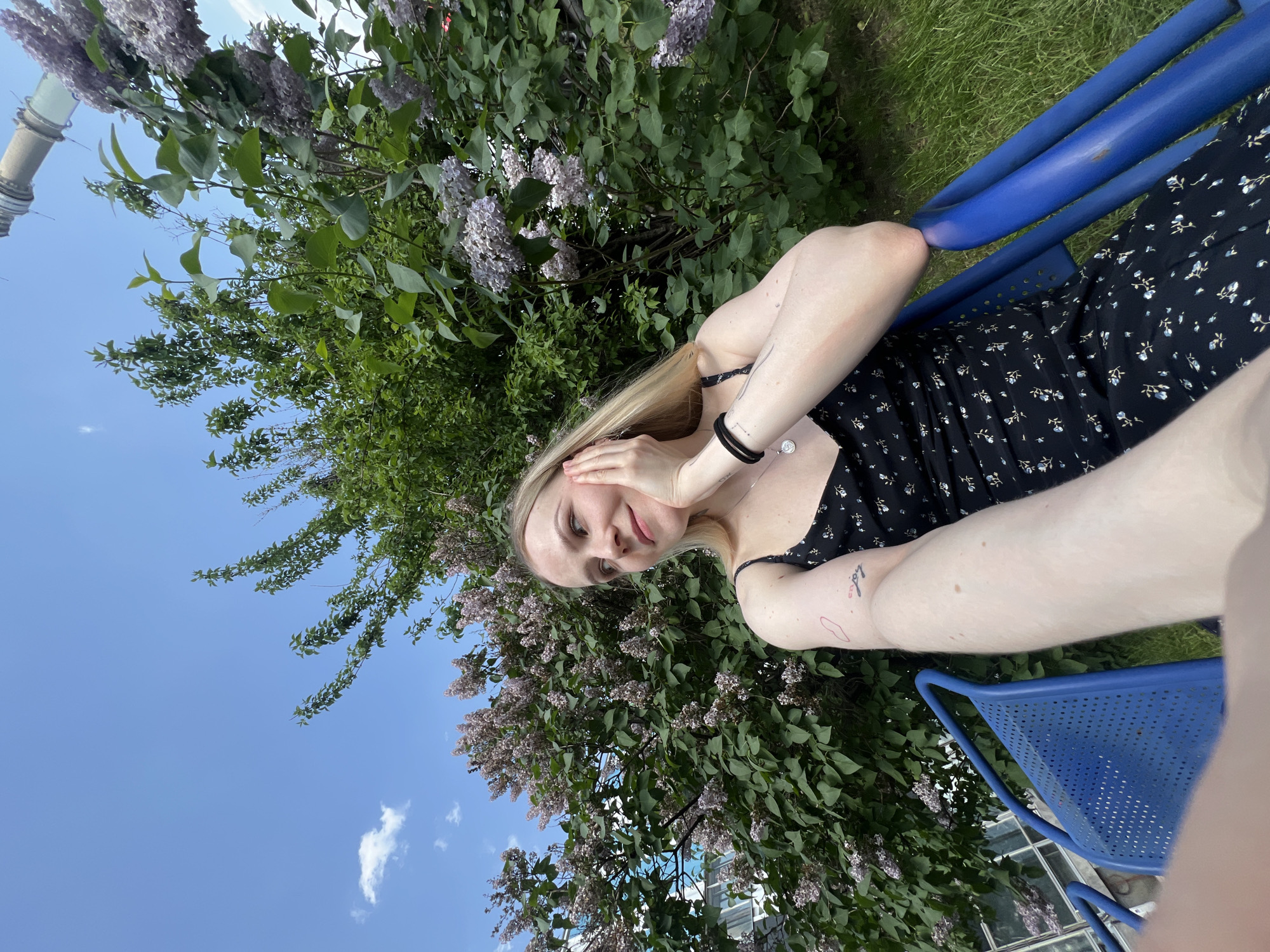 A woman with blonde hair, wearing a black floral dress and a bracelet on her left wrist, sits in a blue metal chair. Her hands are near her face. Lilac bushes surround her, and the sky is visible in the background. In the distance, a structure is visible, suggesting a location in Khimki, Russia.
