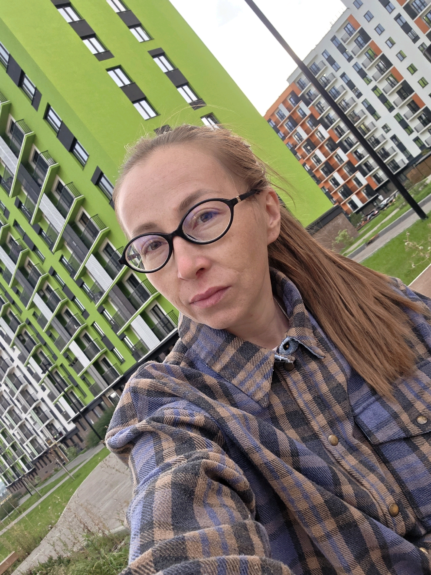 A woman wearing glasses and a plaid shirt is taking a selfie outdoors with a backdrop of modern residential buildings. One building is painted bright green with white window frames, and another building in the background is white with orange and dark gray accents. The woman has long brown hair pulled back into a ponytail and is looking directly at the camera with a neutral expression. The setting appears to be a residential neighborhood, possibly in Pervomaysky, Russia, with well-maintained green spaces and paved pathways visible.
